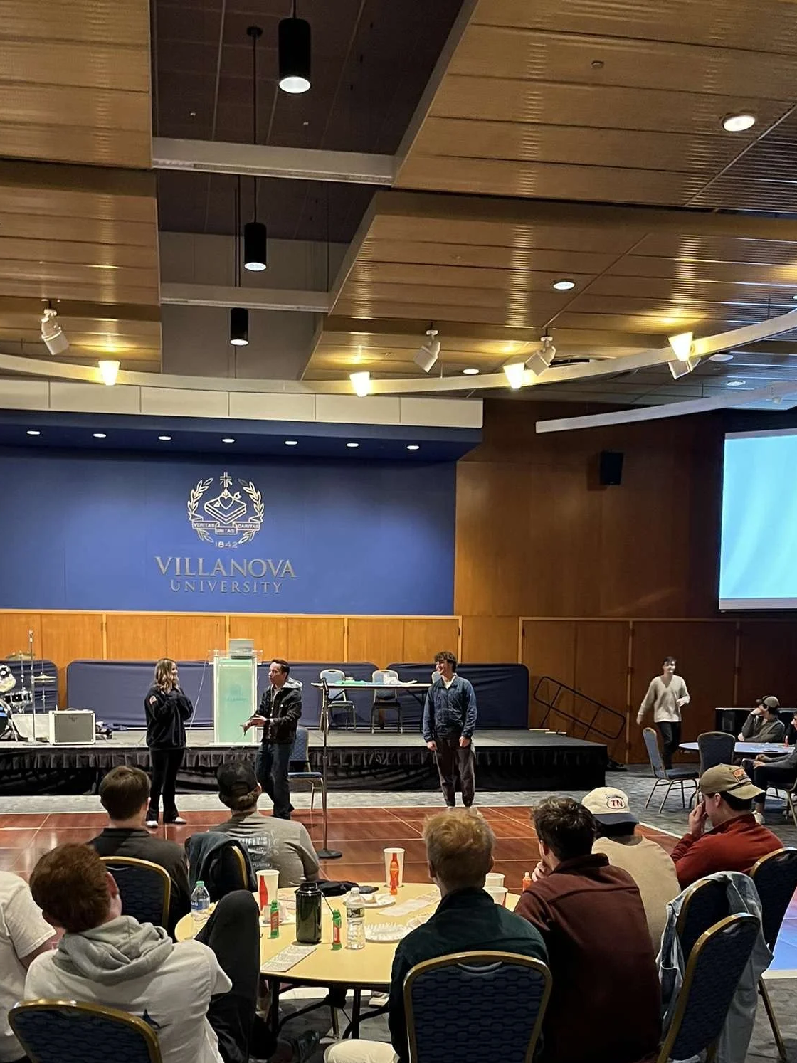 A group of people attending an event at Villanova University, with some individuals speaking on stage and others seated at tables.