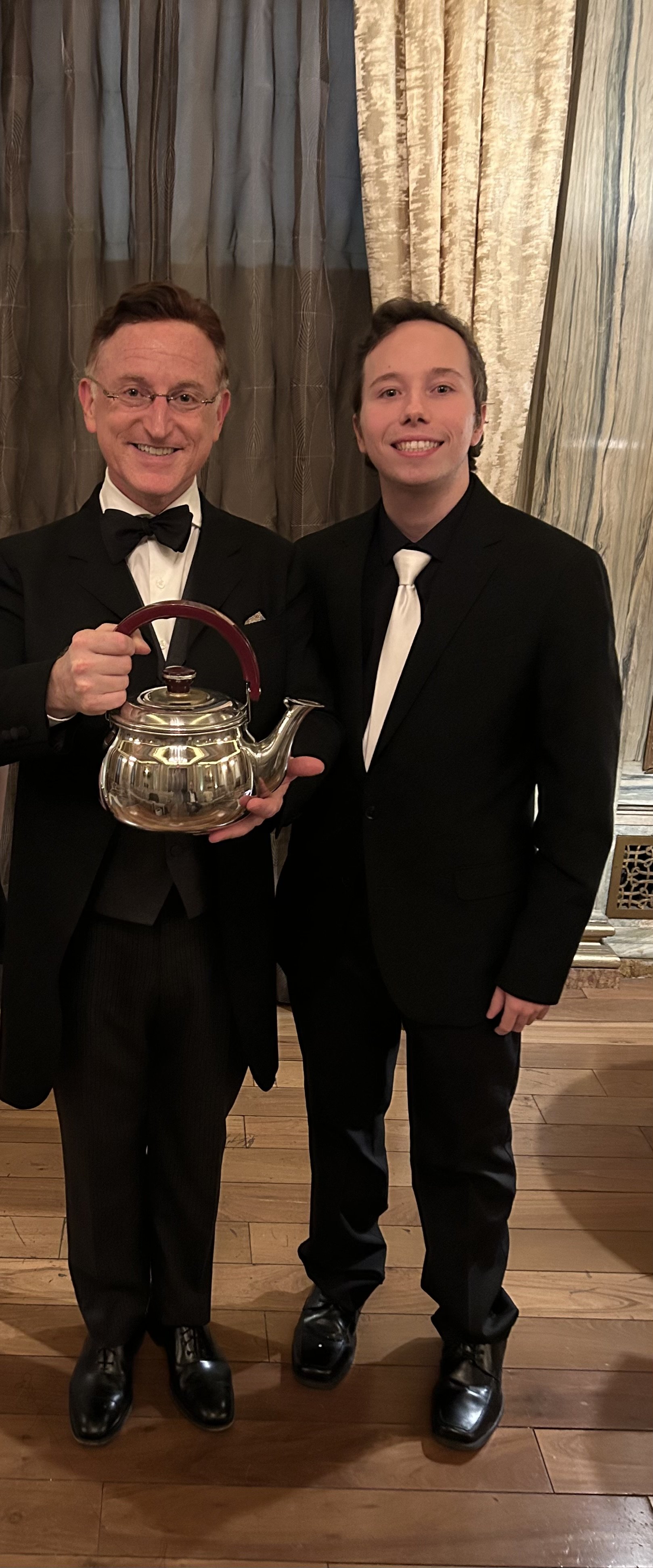 Two men in tuxedos, one holding a silver teapot, standing in a formal indoor setting with curtains and wooden flooring.