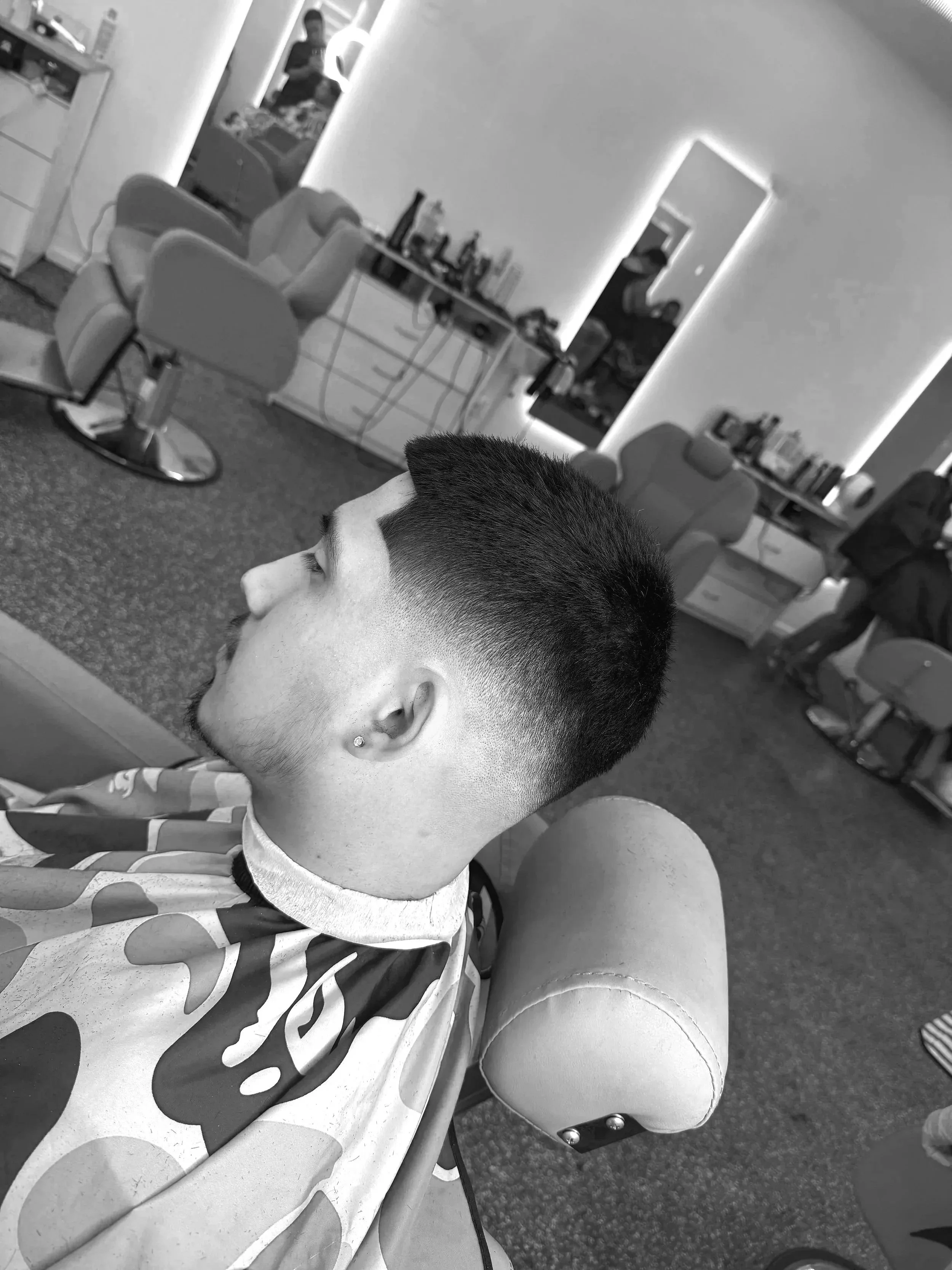 A young man getting a haircut at a barbershop, sitting in a barber chair with a camouflage cape, with barber chairs, mirrors, and hair products visible in the background.