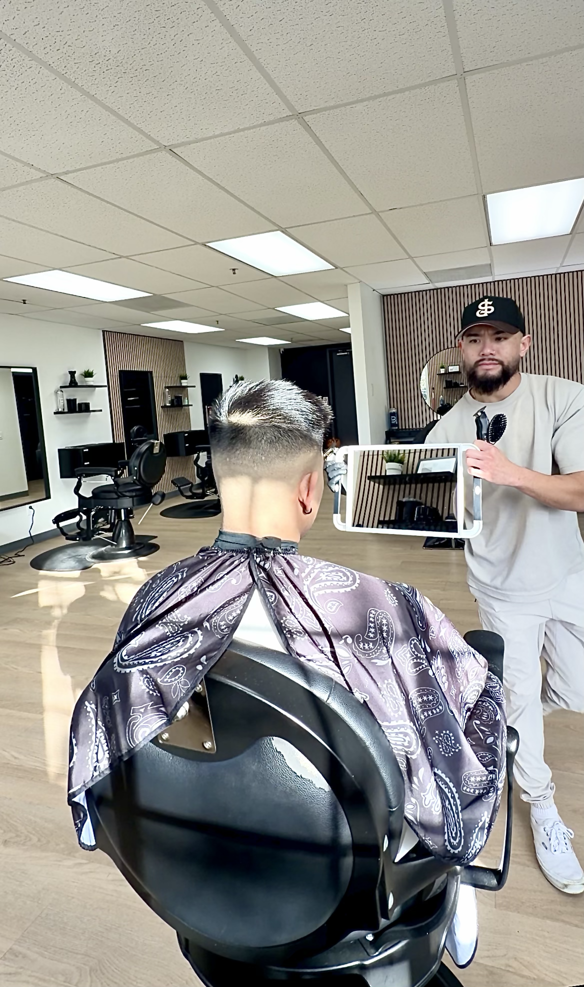 A man with a facial hair and cap holding a mirror and a hairbrush, standing in a hair salon, looking at a young man with a fresh haircut sitting in a salon chair with a patterned cape.