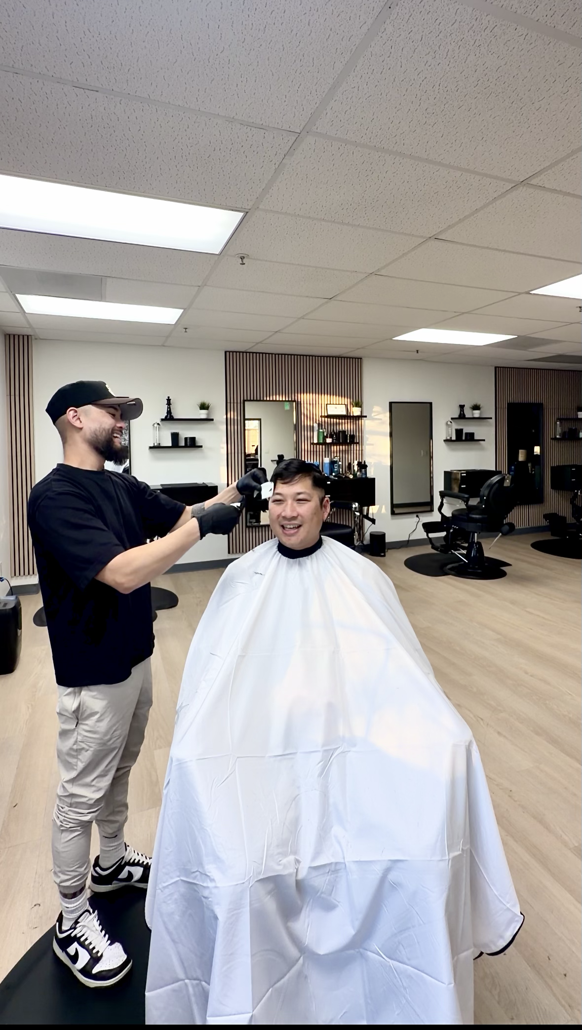 A man gets a haircut from a barber in a salon, smiling and sitting under a white cape in a modern salon setting.