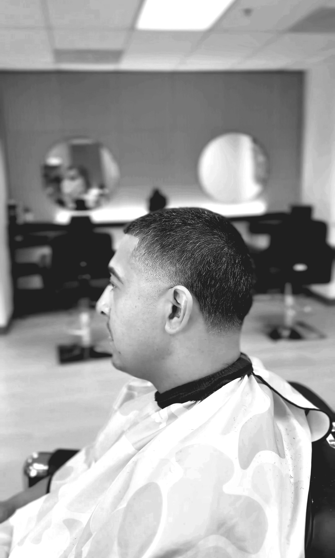 A man with a fresh haircut sitting in a barbershop chair, wearing a barber cape, with a mirror and chairs in the background.