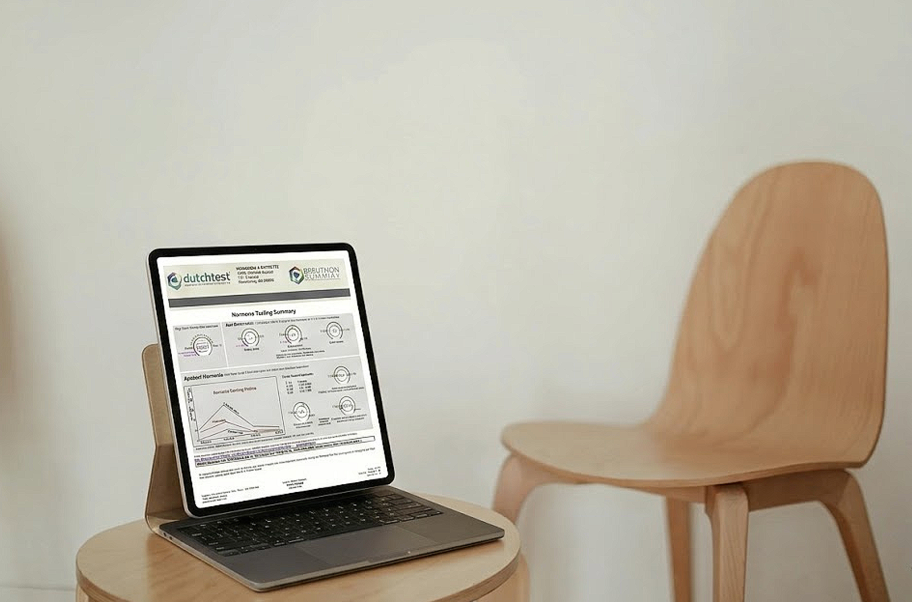Open laptop displaying a report or summary on a light wooden table, with a matching wooden chair beside it, against a plain white wall.