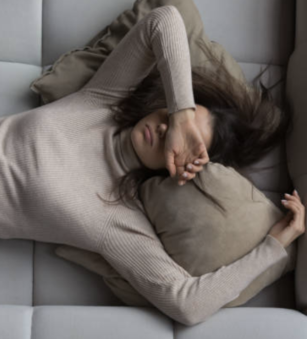 A woman with long dark hair lying on her side on a beige sofa, resting her head on a pillow, and covering her eyes with her arm.