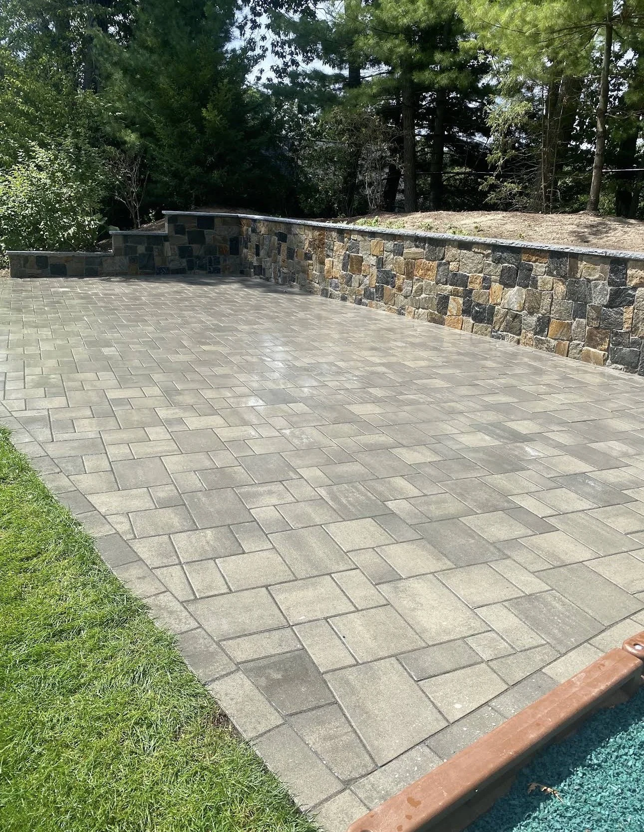 Patio with interlocking concrete pavers, stone retaining wall, grass on the side, trees and forest in the background.