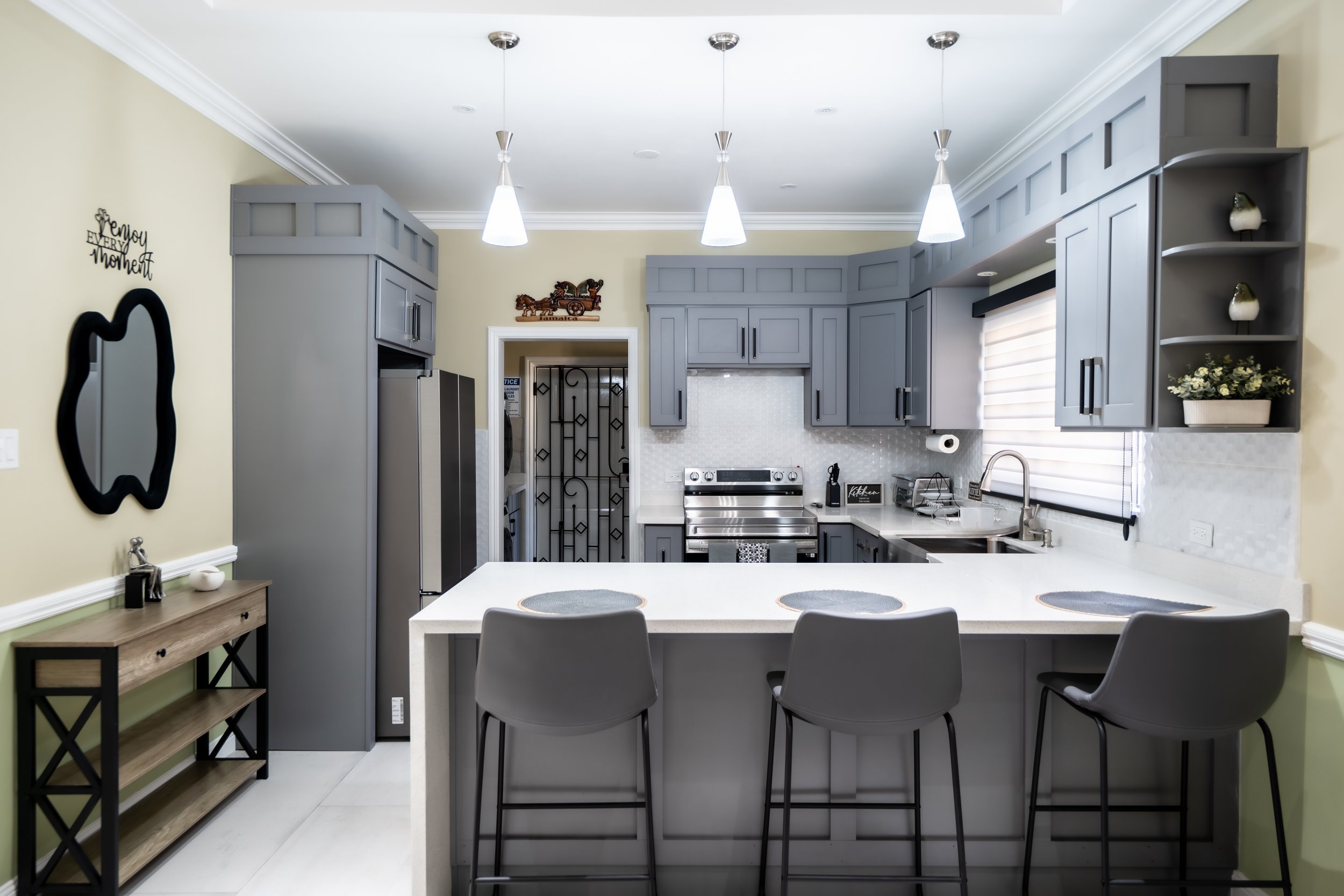 Modern kitchen with grey cabinets, white countertops, and a breakfast bar with three grey chairs. Features include stainless steel appliances, a large window with a shade, decorative items, and hanging pendant lights.