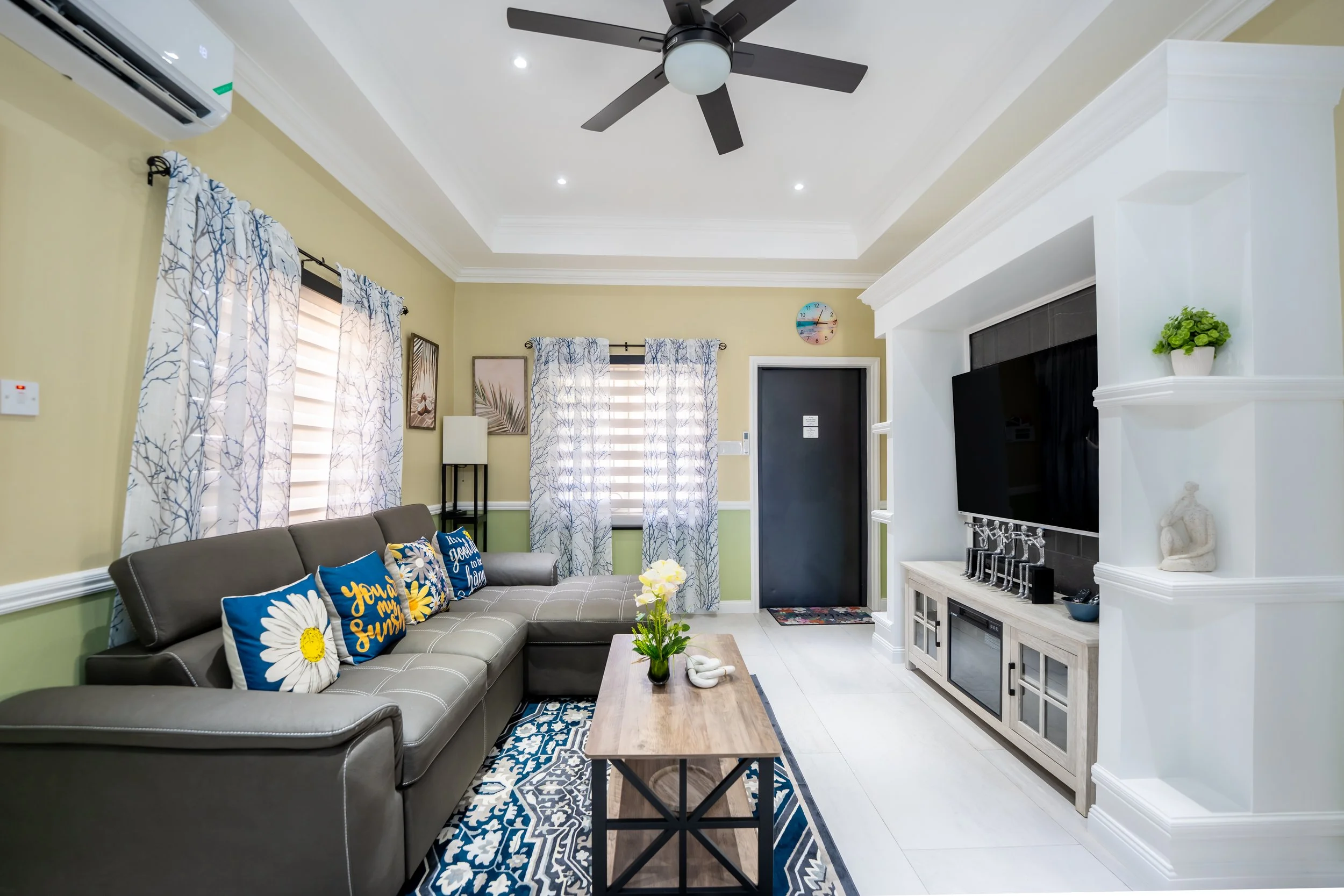 Living room with yellow walls, large gray sectional sofa with colorful pillows, wooden coffee table, white media console with TV, decorative items, and windows with floral curtains, ceiling fan, and light-colored tile floor.