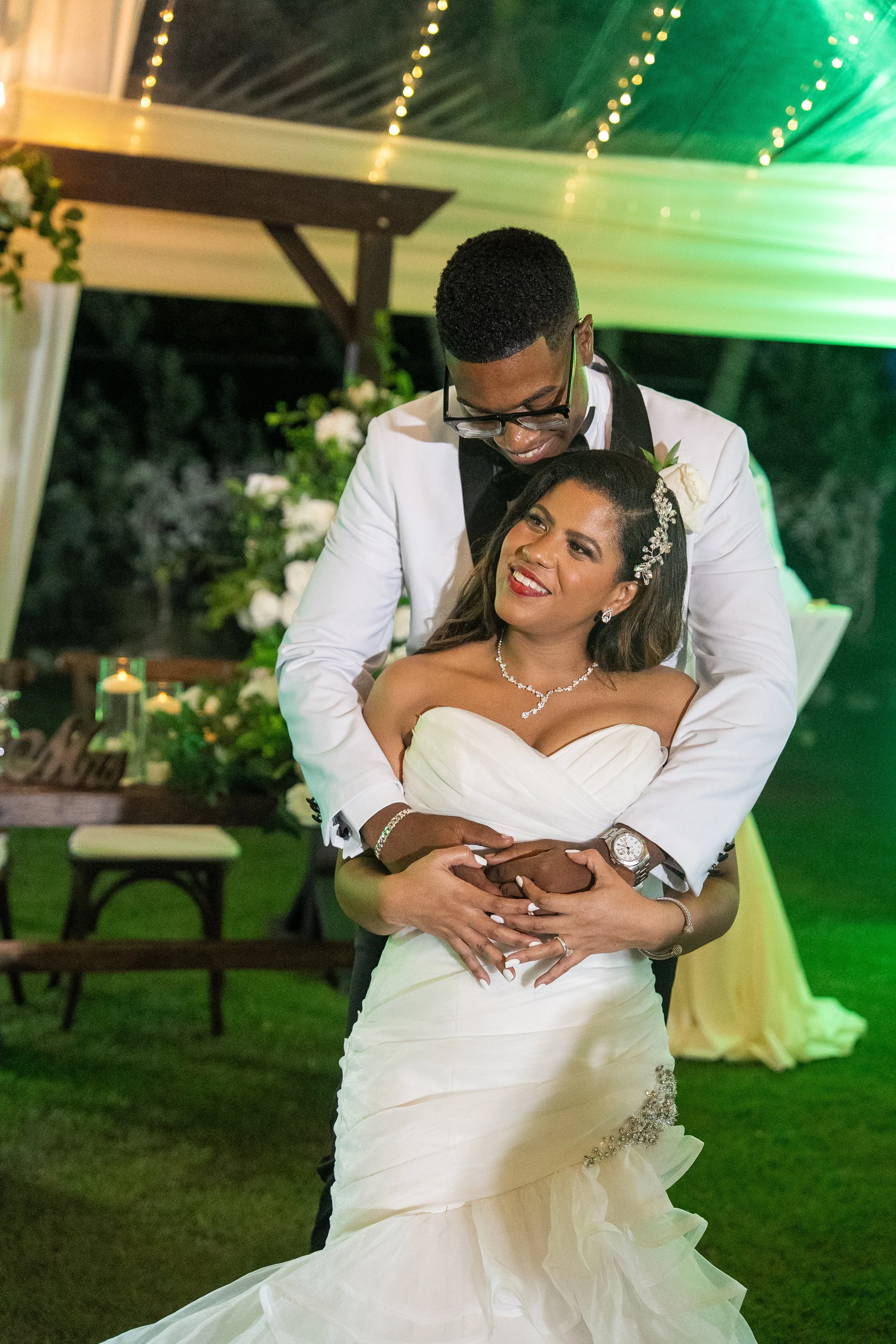 A newlywed couple dancing at their wedding reception, with the groom embracing the bride from behind and both smiling, under string lights at night.