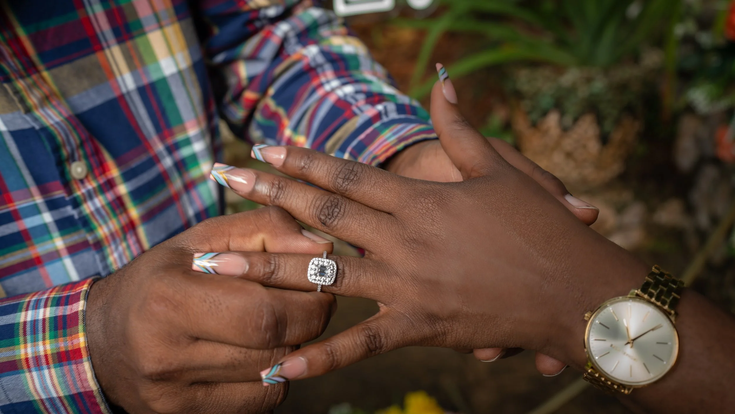 A person is placing a ring on another person's finger. The person receiving the ring is wearing a gold watch, and the person giving it is dressed in a colorful checkered shirt. The photograph is taken outdoors with blurred plants in the background.