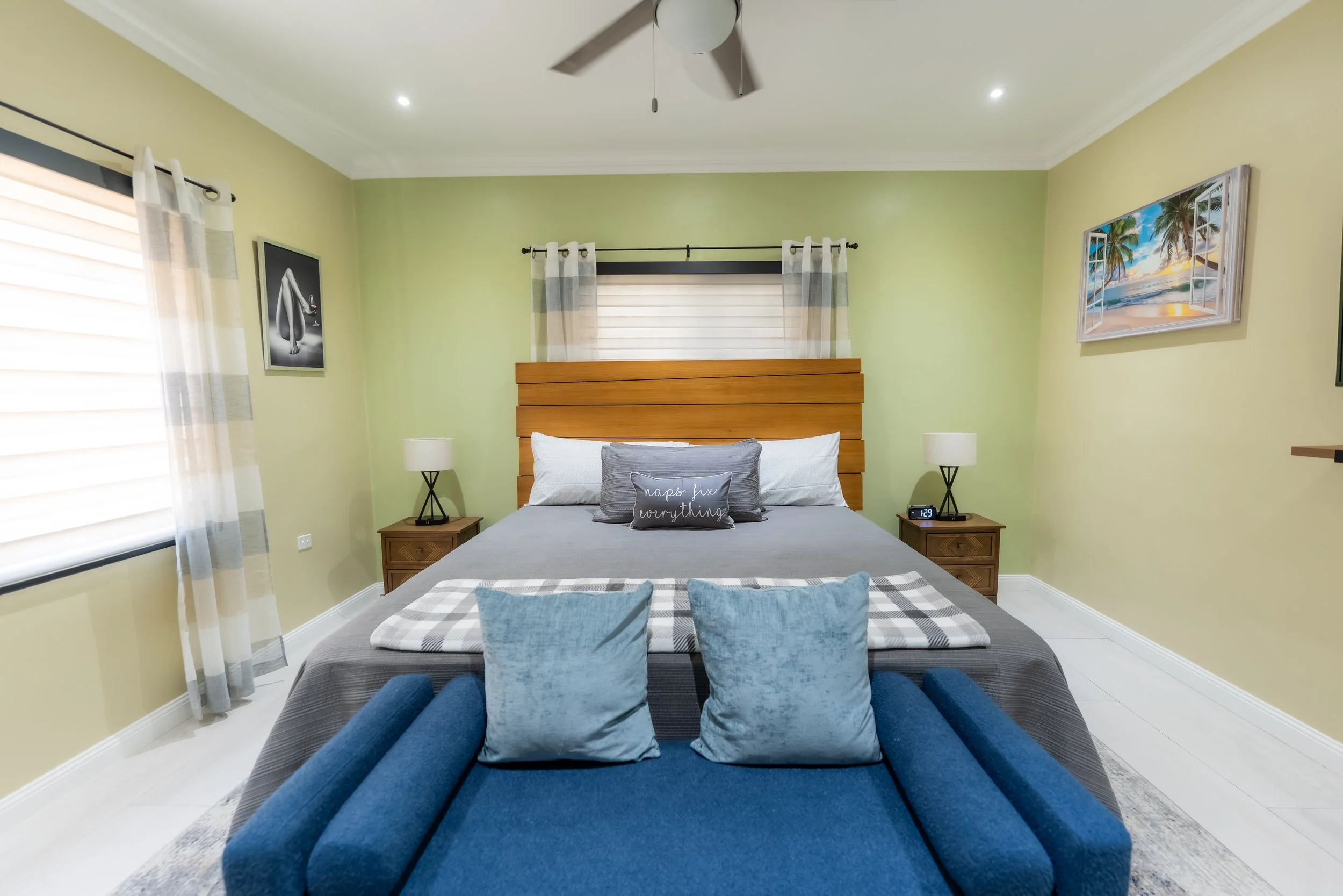 A bedroom with a bed, two nightstands with lamps, and wall art, featuring pale green walls, white curtains on windows, and a wooden headboard.