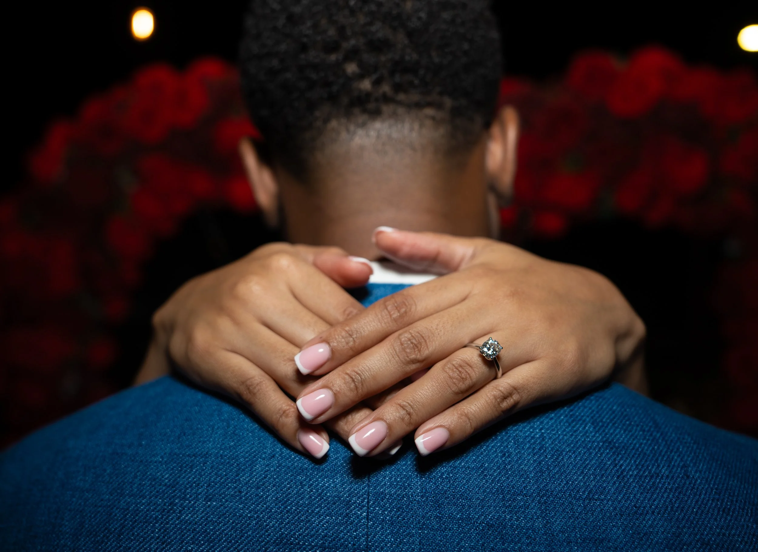 A woman with manicured nails and an engagement ring hugs a man in a blue suit, with a backdrop of red roses.