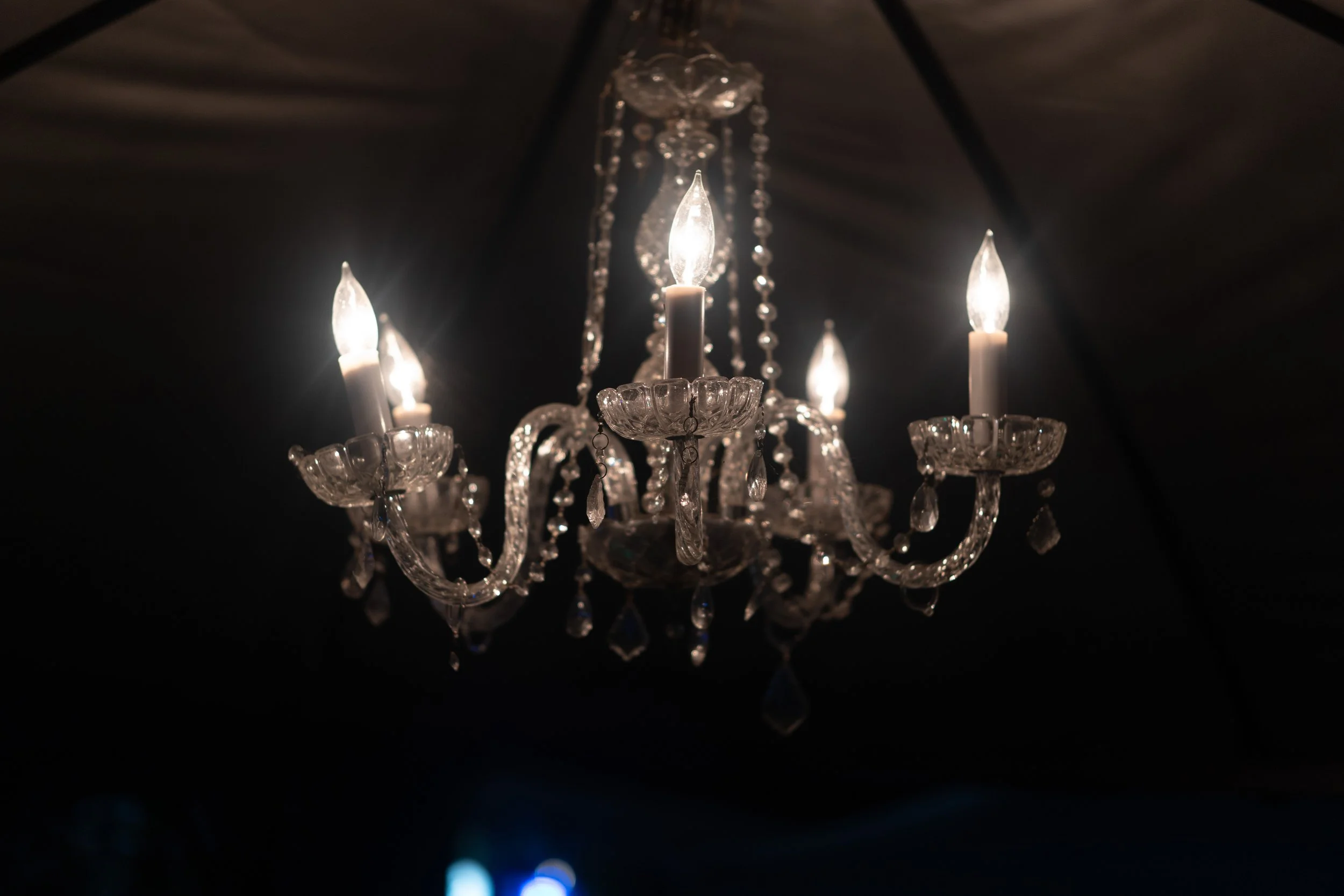 A chandelier with multiple candle-shaped lights and hanging crystal decorations against a dark background.