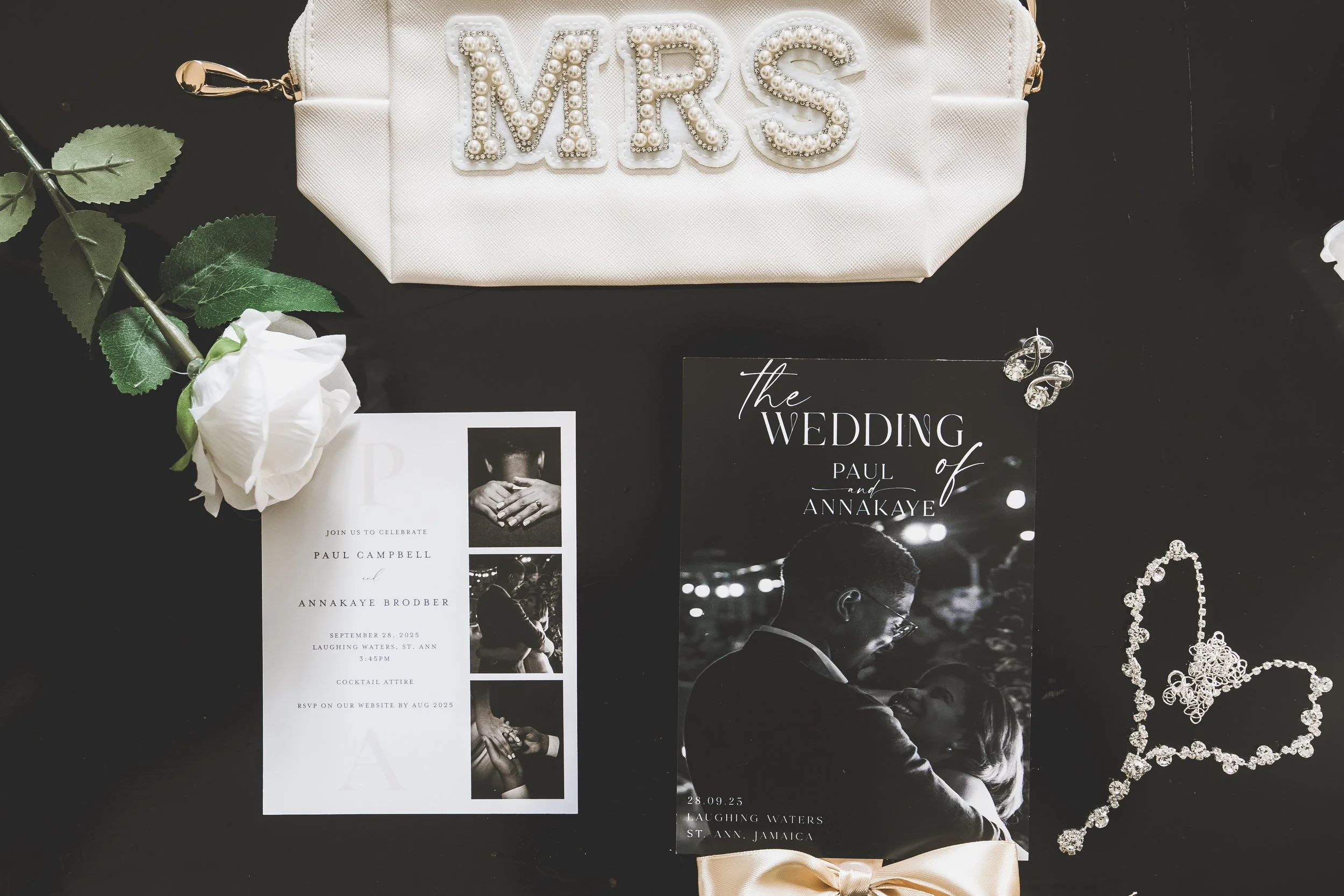Wedding invitation, a white rose, pearl-embellished white clutch with 'MRS' spelled out in pearl letters, diamond necklace, and diamond earrings displayed on a black surface.