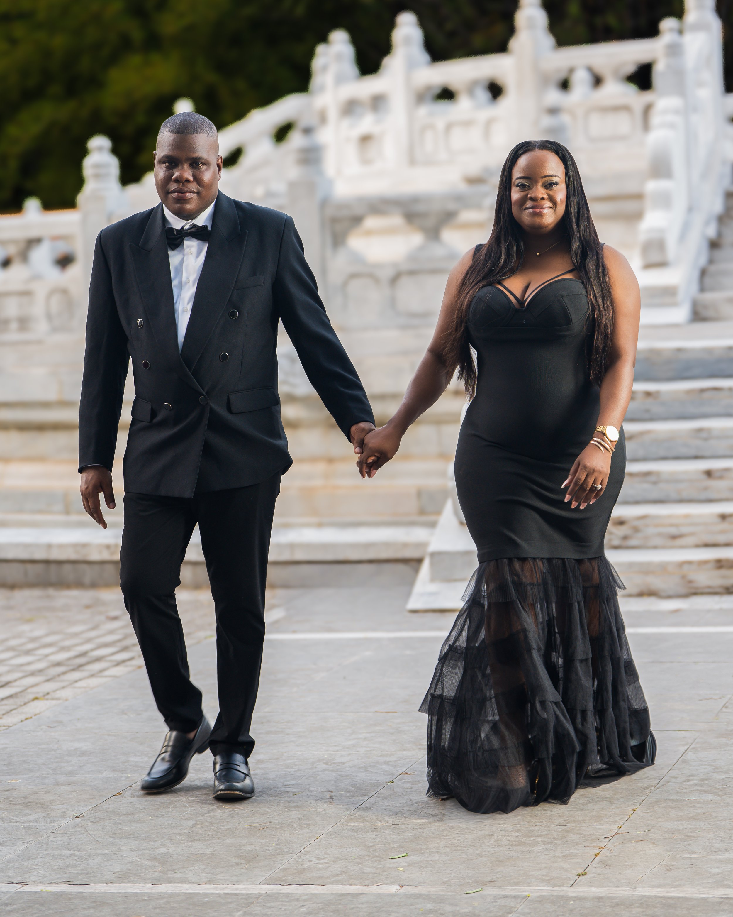 A couple holding hands, dressed elegantly, standing in front of a white stone staircase and railing. The man is wearing a black tuxedo with a bow tie, and the woman is wearing a long, black dress with lace details at the bottom.
