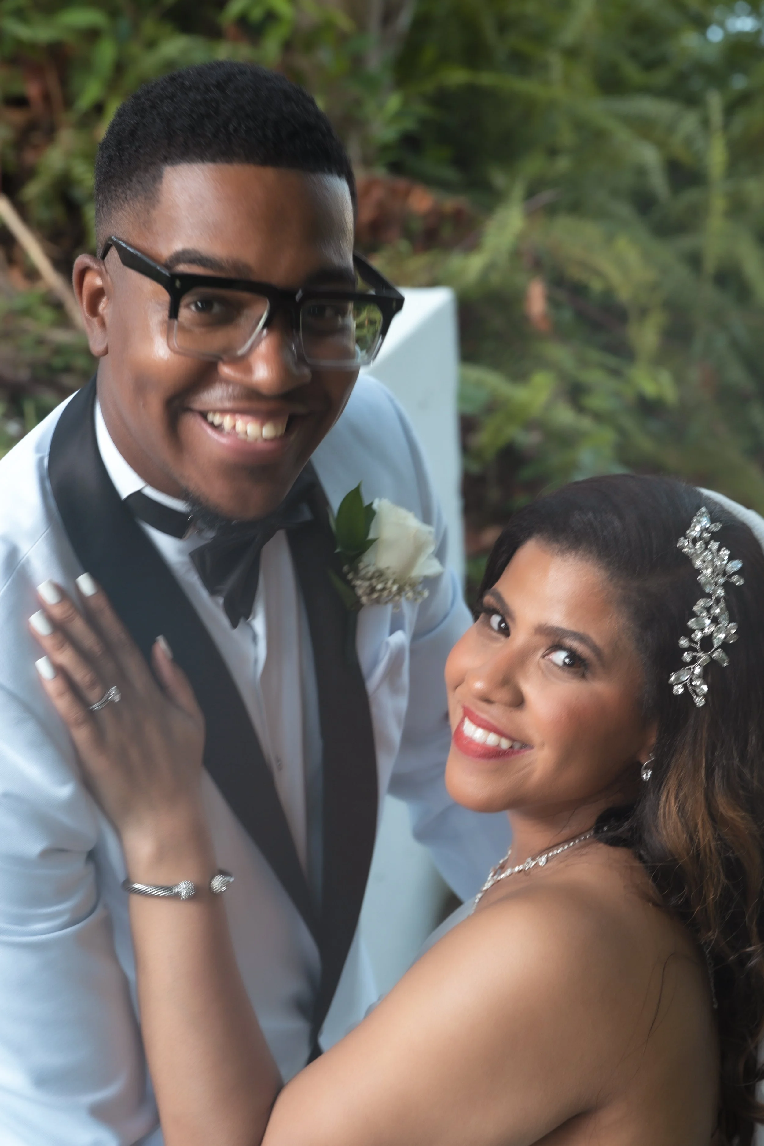 A happy couple dressed in formal wedding attire, with the man wearing glasses, a white tuxedo with black accents, and a boutonniere, and the woman wearing a strapless dress with a crystal hairpiece, smiling at the camera outdoors with greenery in the