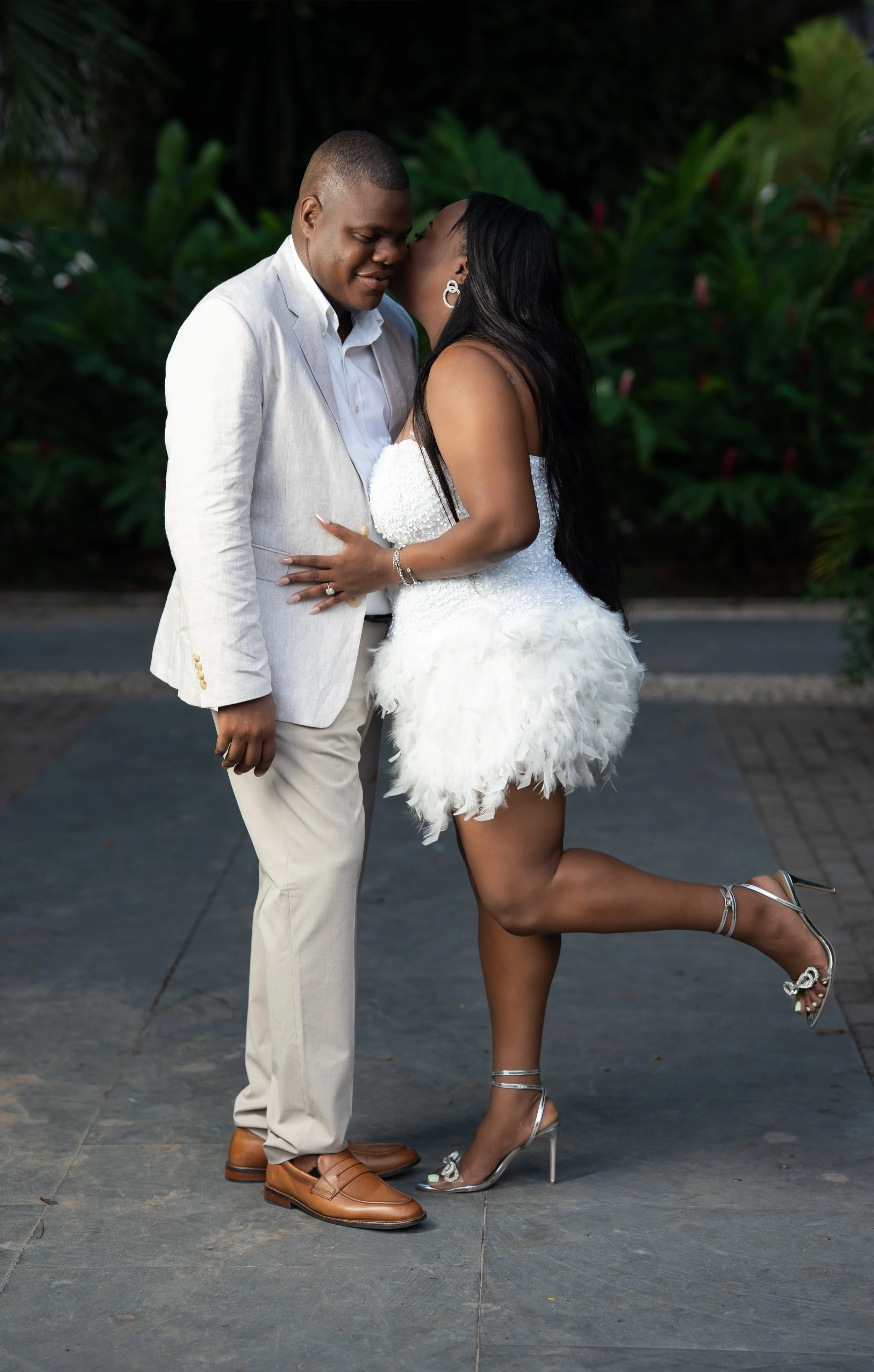 A man and a woman are standing close together outdoors, with the woman whispering into the man's ear. The man is wearing a light-colored suit and dress shoes, and the woman is wearing a white strapless dress with a feathered skirt and high heels. Lus