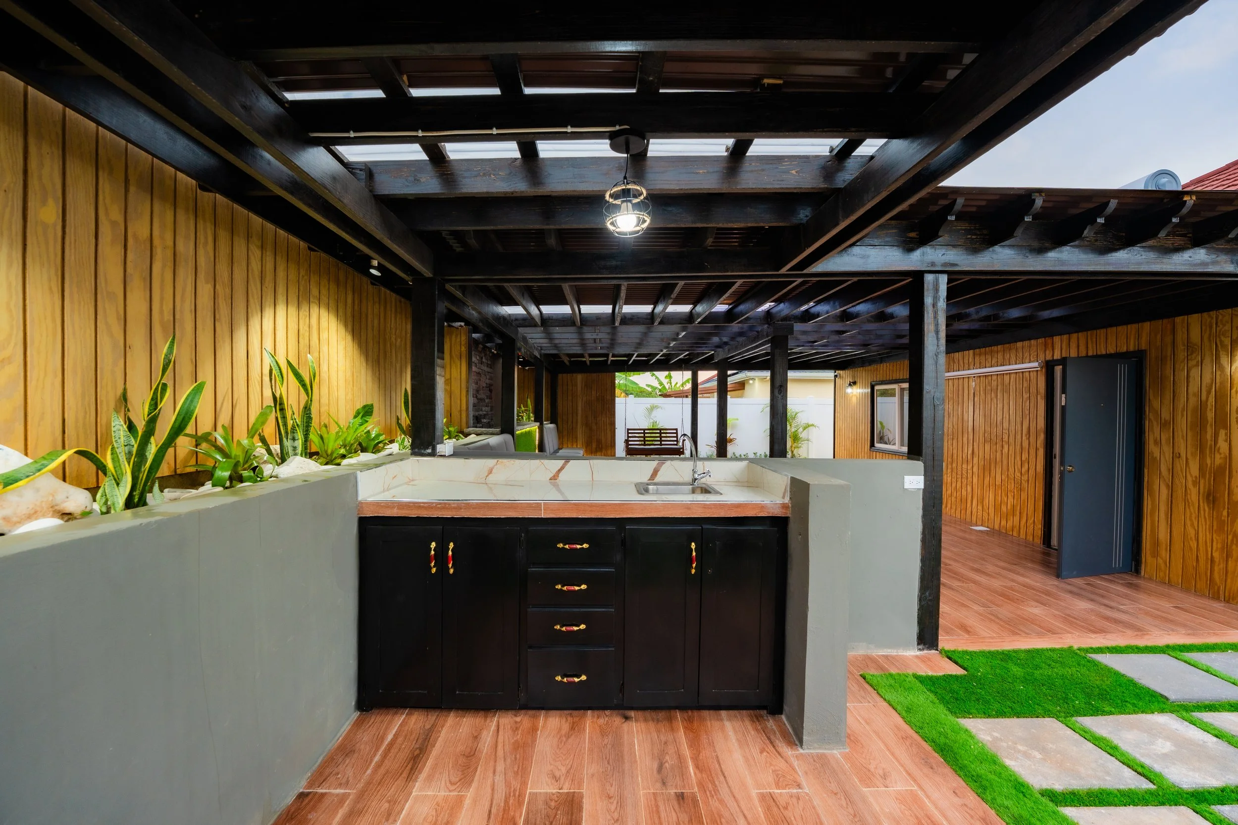 Outdoor kitchen area with black cabinets, marble countertop, wooden flooring, green plants along a wooden wall, and a seating area in the background under a wooden pergola.
