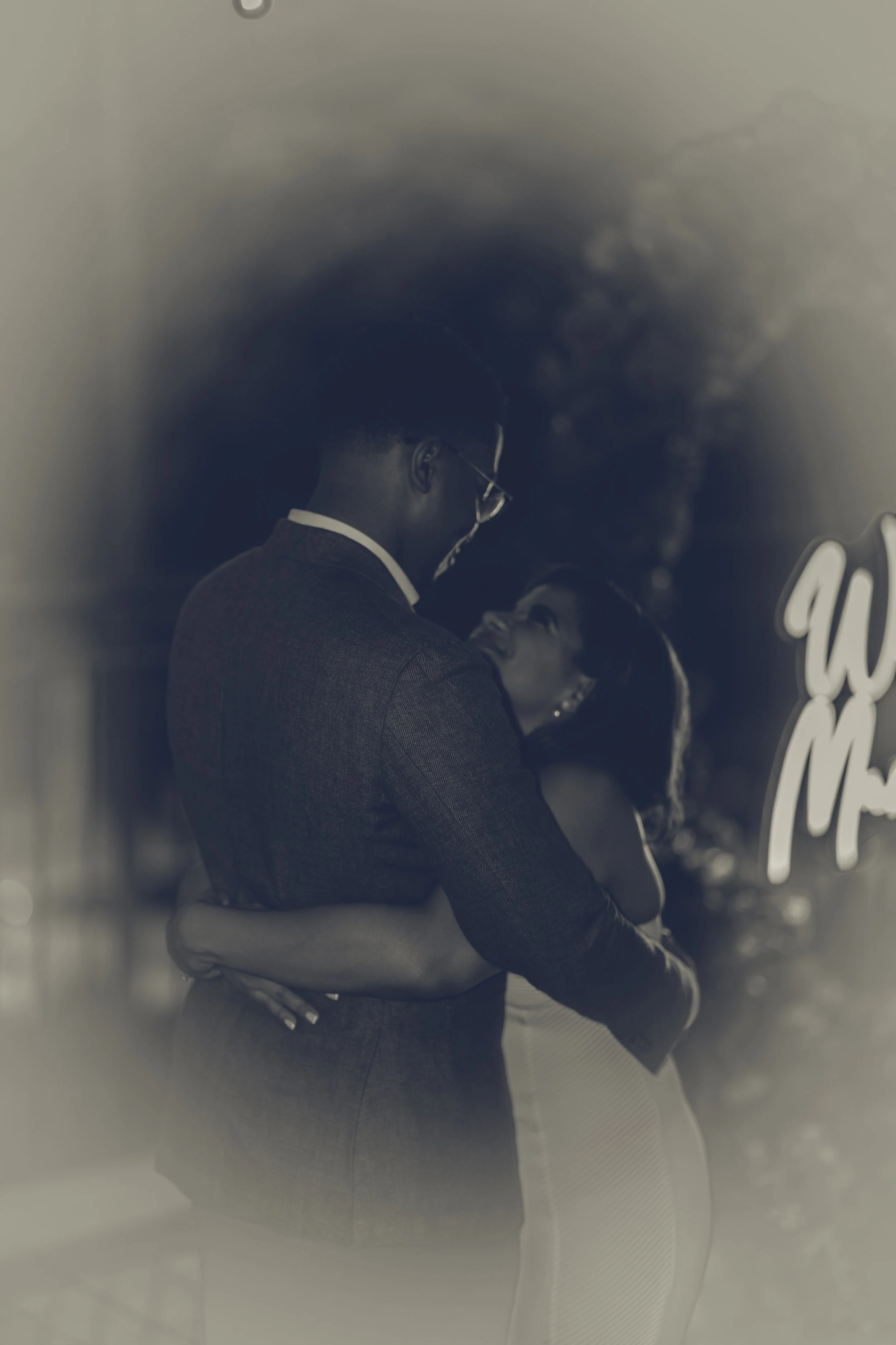 A couple is embracing and smiling at each other, with the man wearing glasses and a suit and the woman wearing a dress, in a romantic moment.