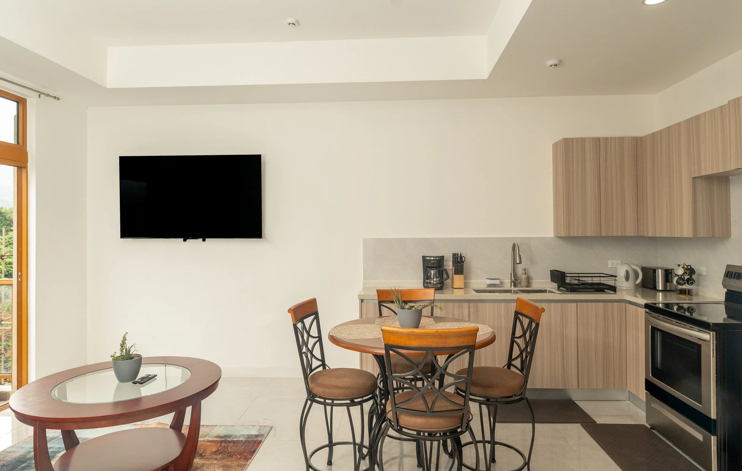 Open kitchen and dining area with a wall-mounted flat-screen TV, round dining table with four chairs, bar-style coffee table, kitchen counter with appliances, and a sliding glass door leading outside.