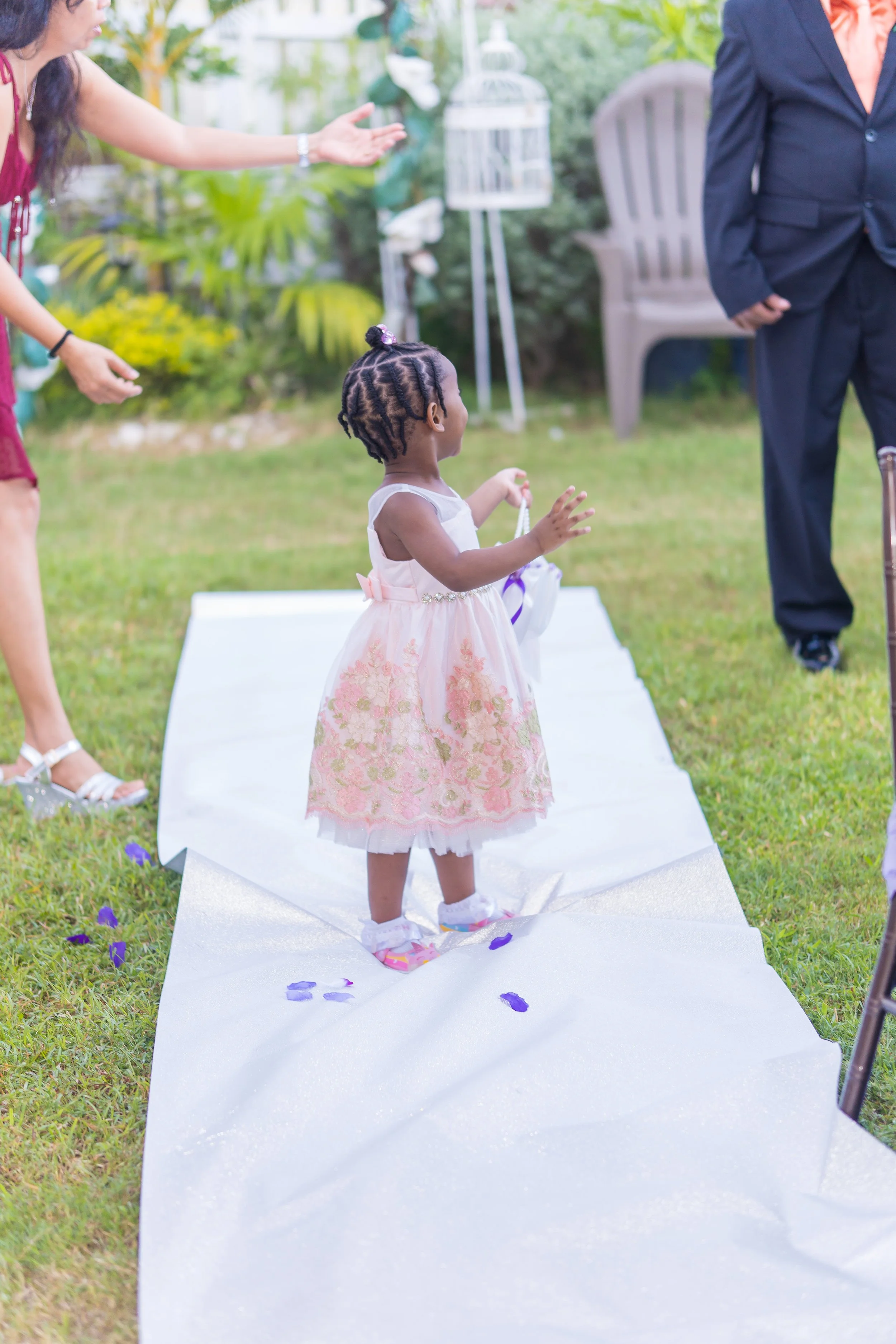 A young girl in a pink floral dress stands on a white aisle runner outdoors, surrounded by adults, with one adult reaching out towards her. There are purple flower petals on the grass and the aisle, with greenery and garden decor in the background.