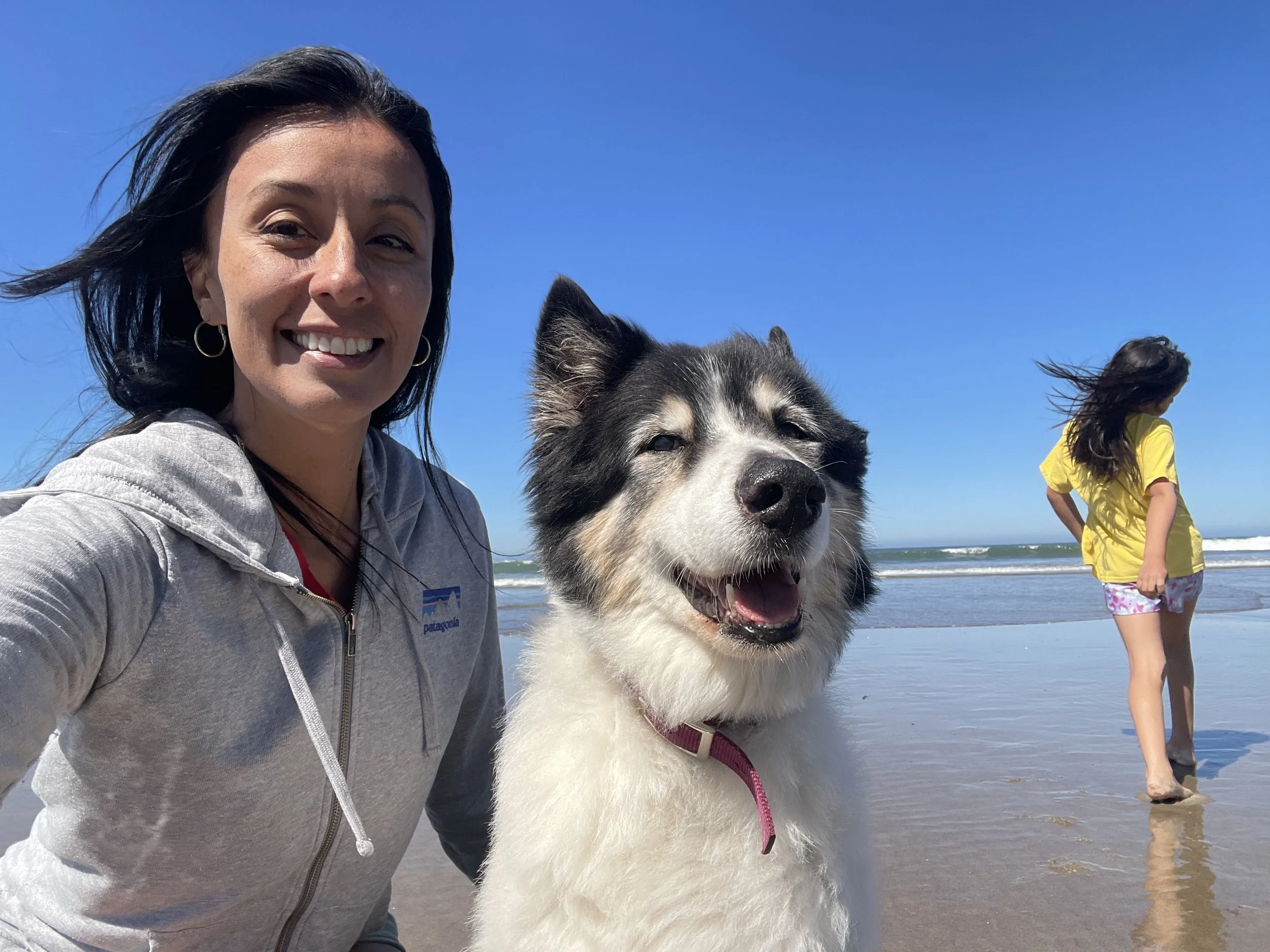 Our fearless leader, Angie Biornstad-Merchan, smiling while sitting on the beach at Rockaway in Oregon with her Akido Shephard mix and her daughter in the background. Angie loves the beach and very much misses her dog, Blake, who passed in 2026.