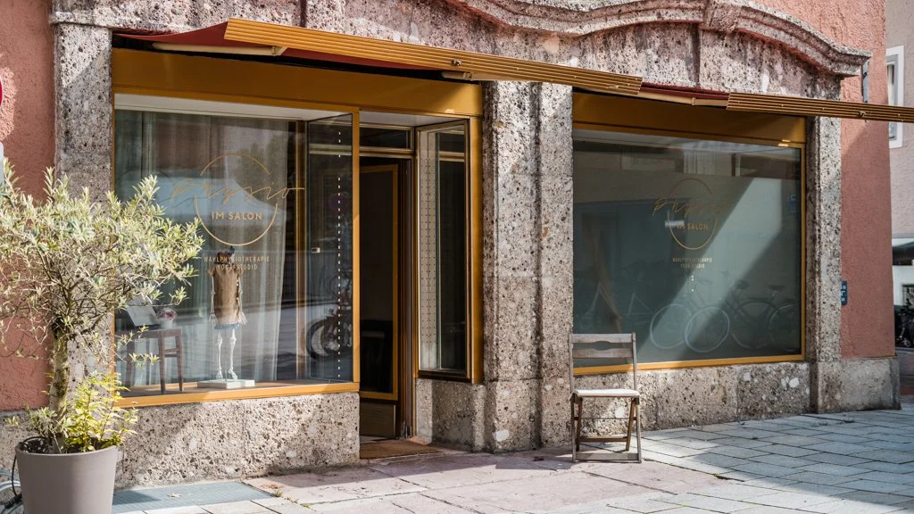 Außenansicht der Praxis "Physio im Salon", in einem ehemaligen Frisiersalon, mit großen Fenstern und einem Sessel und einer Pflanze davor.