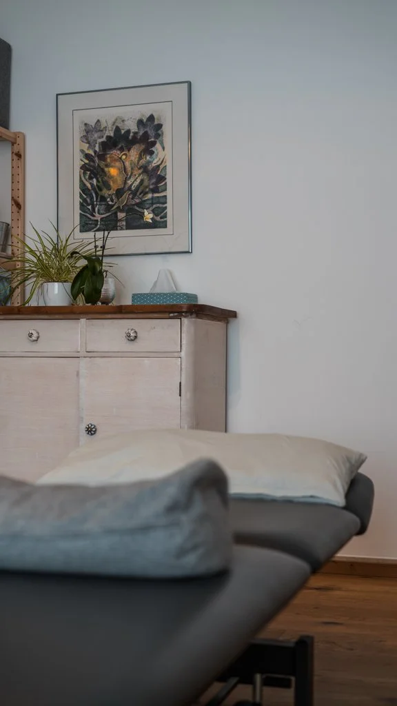 Ein Blick auf einen Praxis-Raum mit Therapie-Liege im Vordergrund, im Hintergrund sind ein Schrank, einer Zimmerpflanze und ein Wandbild sichtbar.