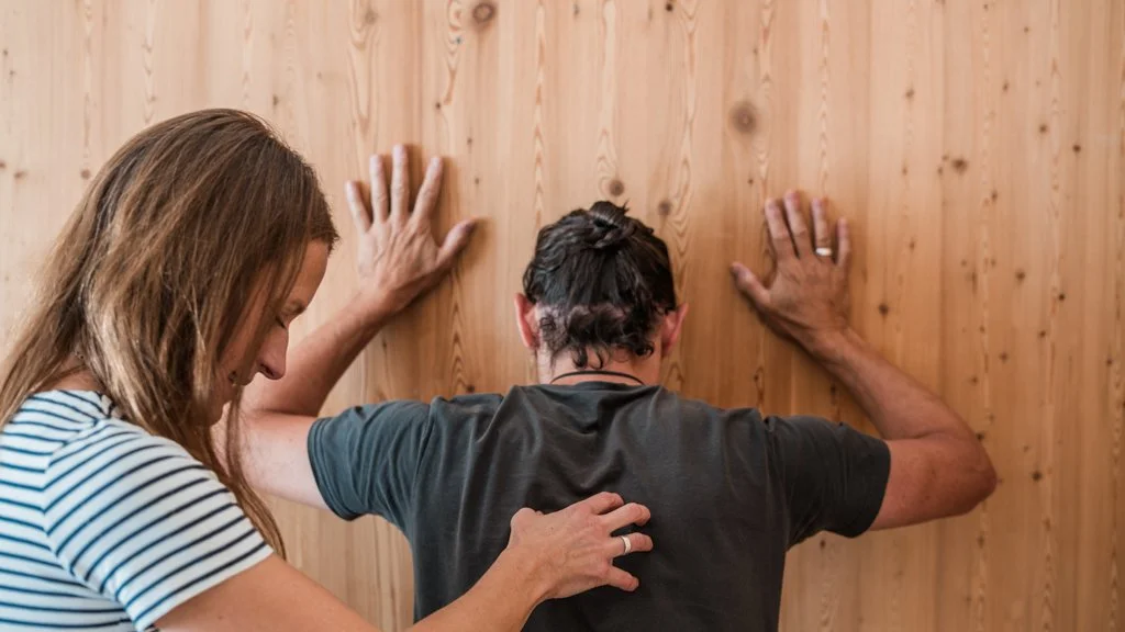 Eine Frau behandelt den Rücken eines Mannes, der gegen eine Holzwand lehnt, mit dem Rücken zur Kamera.