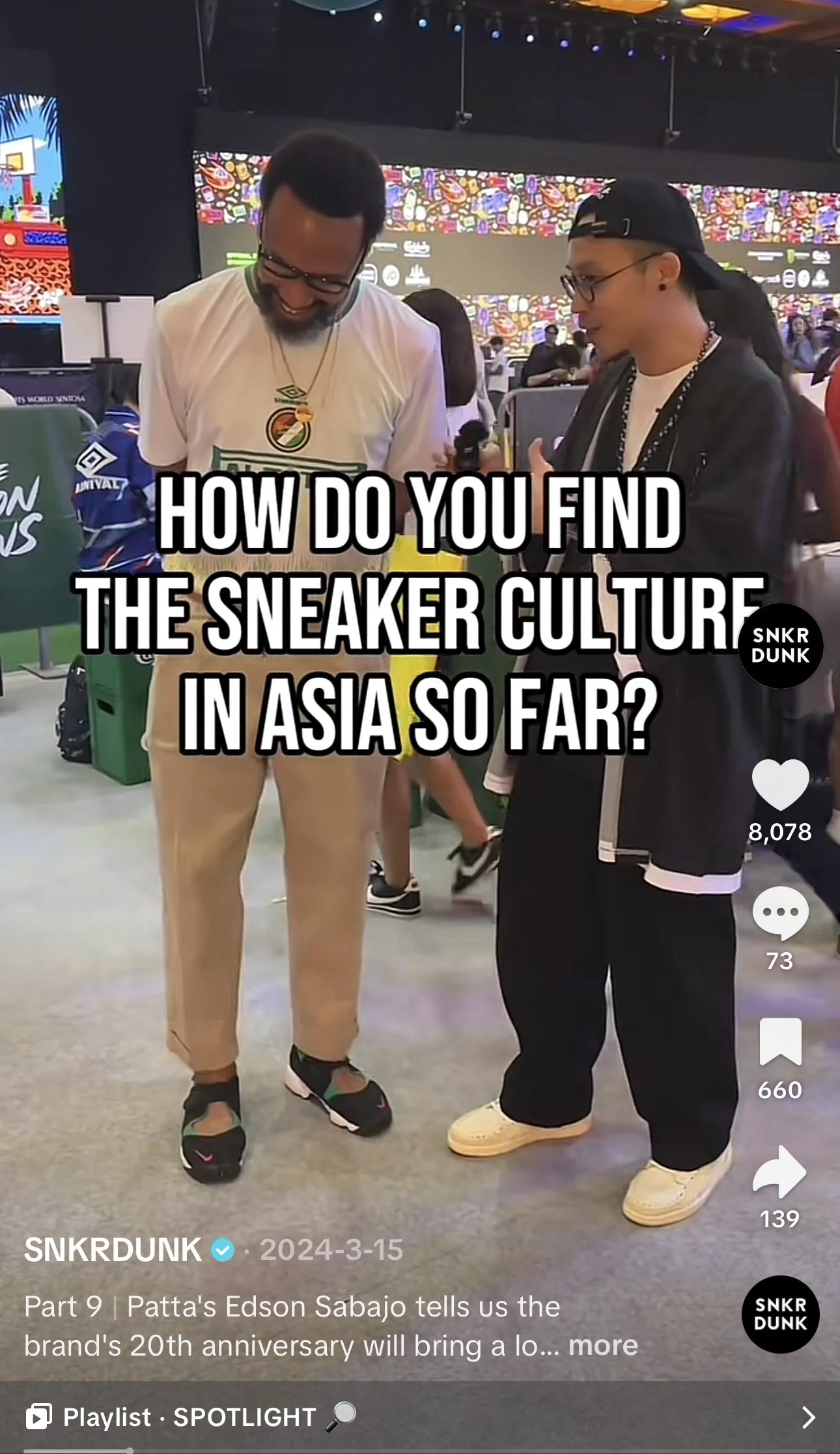 Two men are talking at an indoor event, one wearing a white T-shirt and beige pants, the other in black clothing with a black cap, both smiling. There are colorful screens and other attendees in the background.