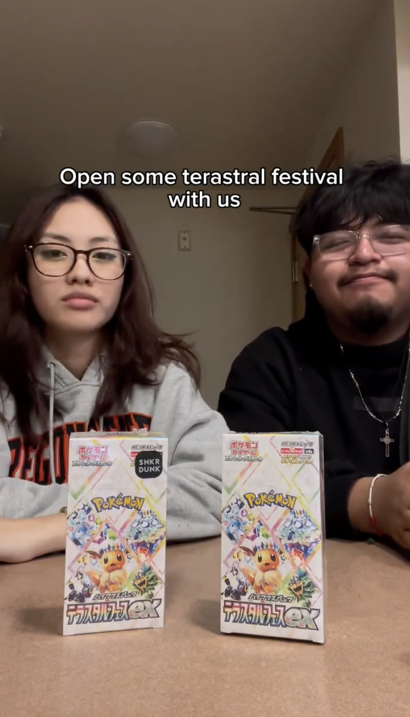 Two people sitting at a table with two Pokémon-themed candy boxes in front of them. The person on the left has glasses and long brown hair, wearing a gray hoodie. The person on the right has glasses, dark hair, and a beard, wearing a black shirt and a cross necklace. The background is an indoor room with a beige wall and a ceiling light. There are subtitles that say, 'Open some terastral festival with us.'