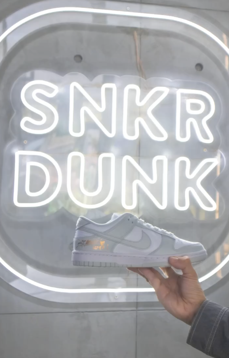 Hand holding a white Nike sneaker in front of a neon sign that says "SNKR DUNK."