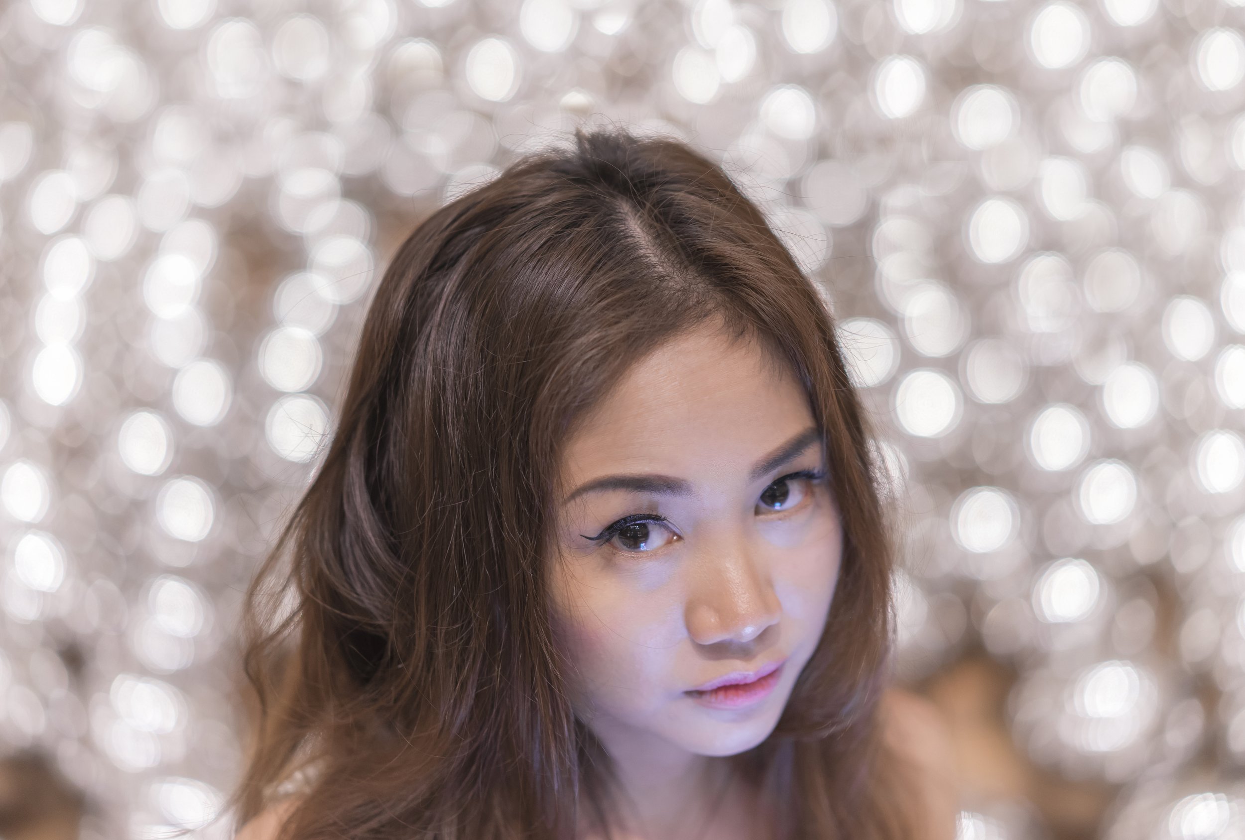 A woman with long brown hair and winged eyeliner looking slightly upward, with a blurred shiny, silver background.