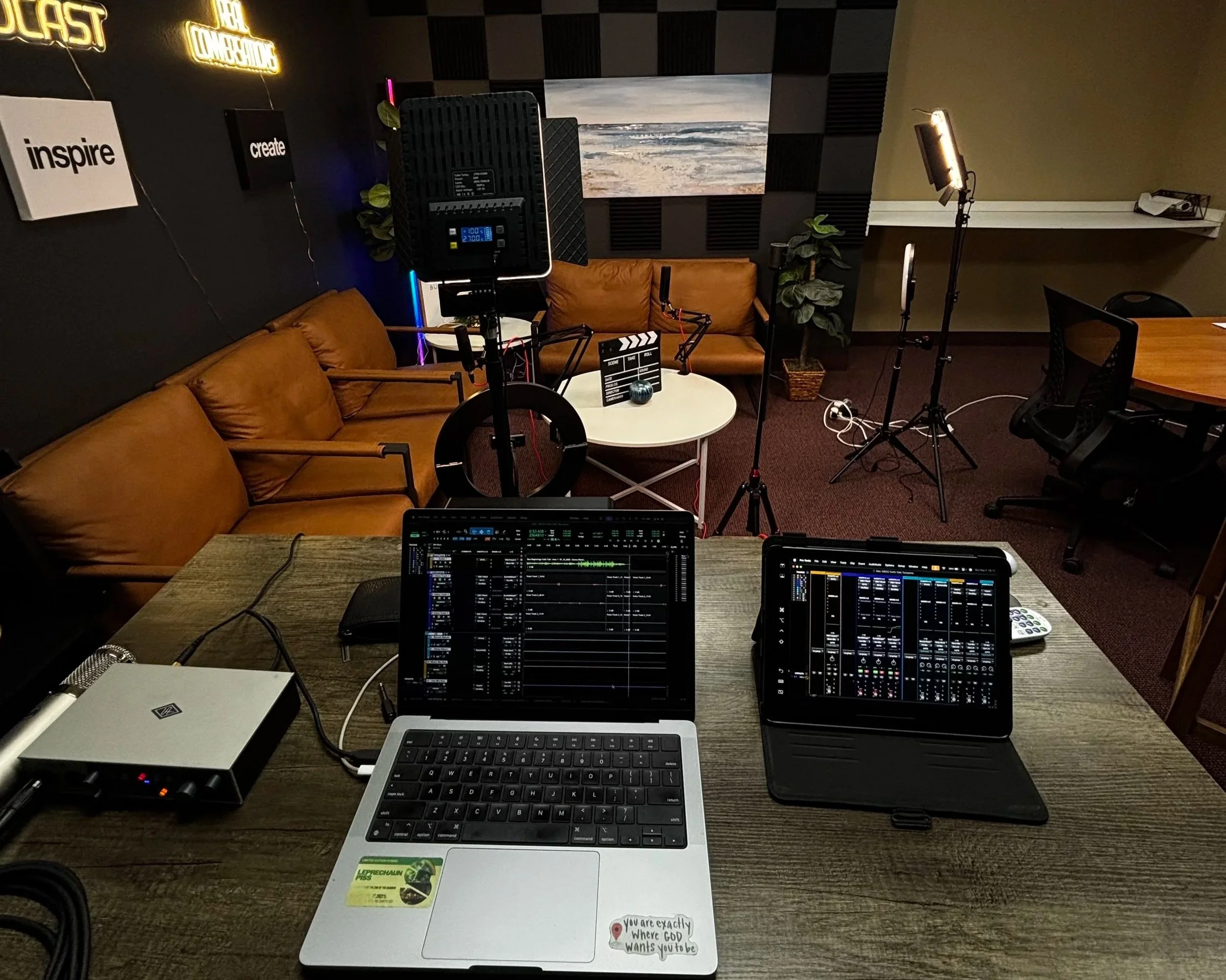 A recording studio setup with a table, laptop, and digital controller, microphones, studio lights, couches, and decorations.