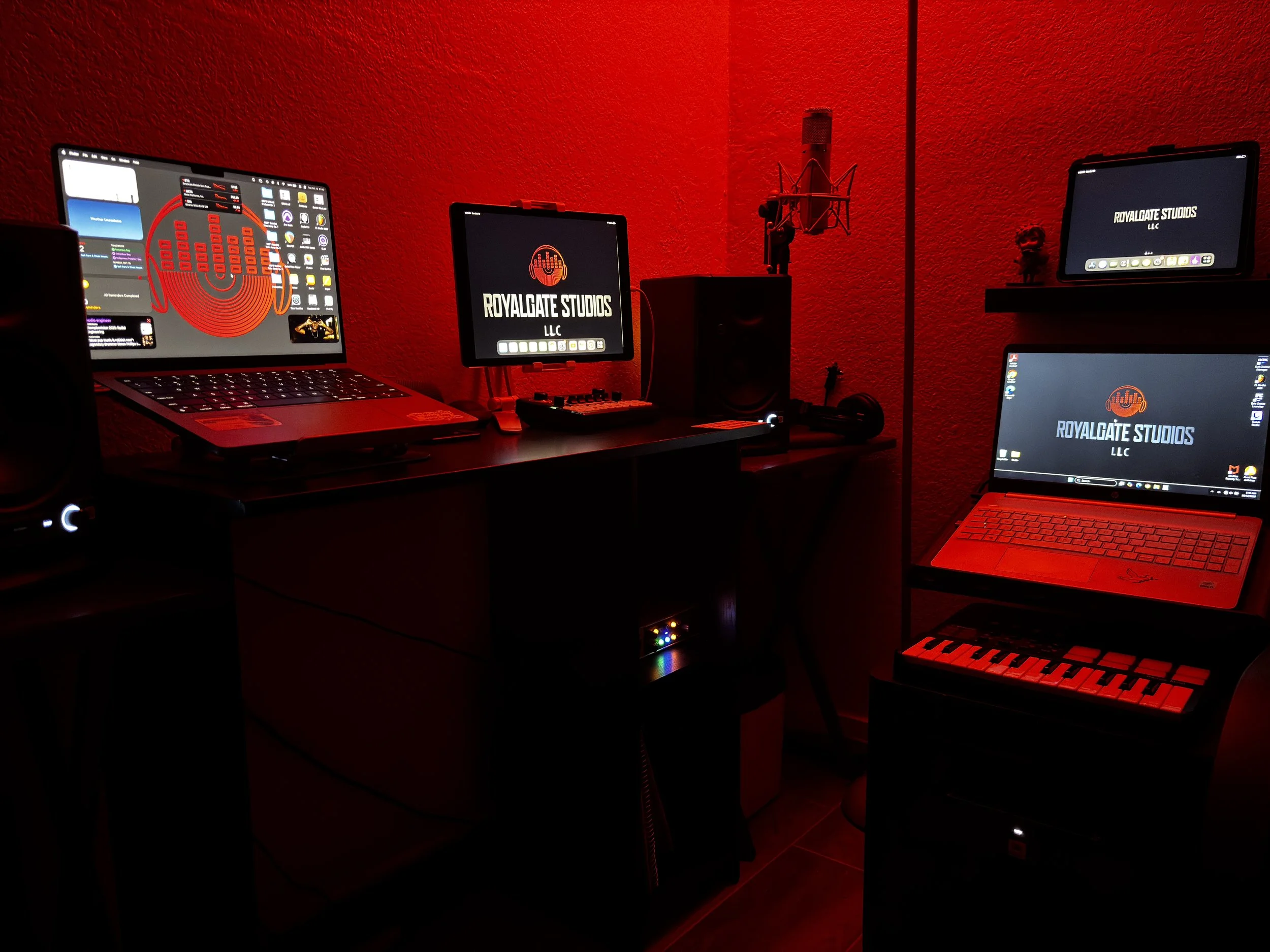 A music production studio with multiple computer monitors, keyboards, a microphone, and studio speakers, all illuminated by red lighting. The screens display a logo that reads "Royalgate Studios LLC."