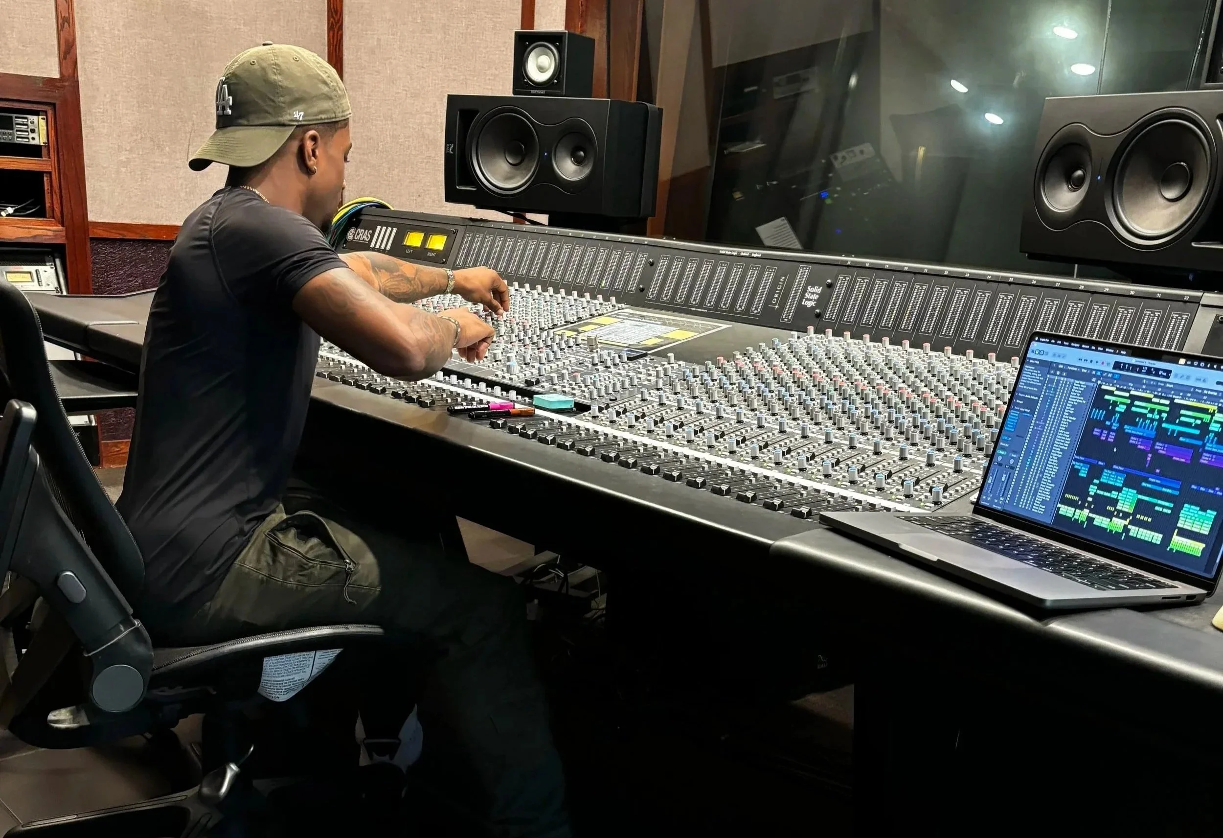 A man wearing a green cap and black t-shirt sitting in front of a large audio mixing console in a recording studio, with a laptop displaying music production software and studio speakers in the background.