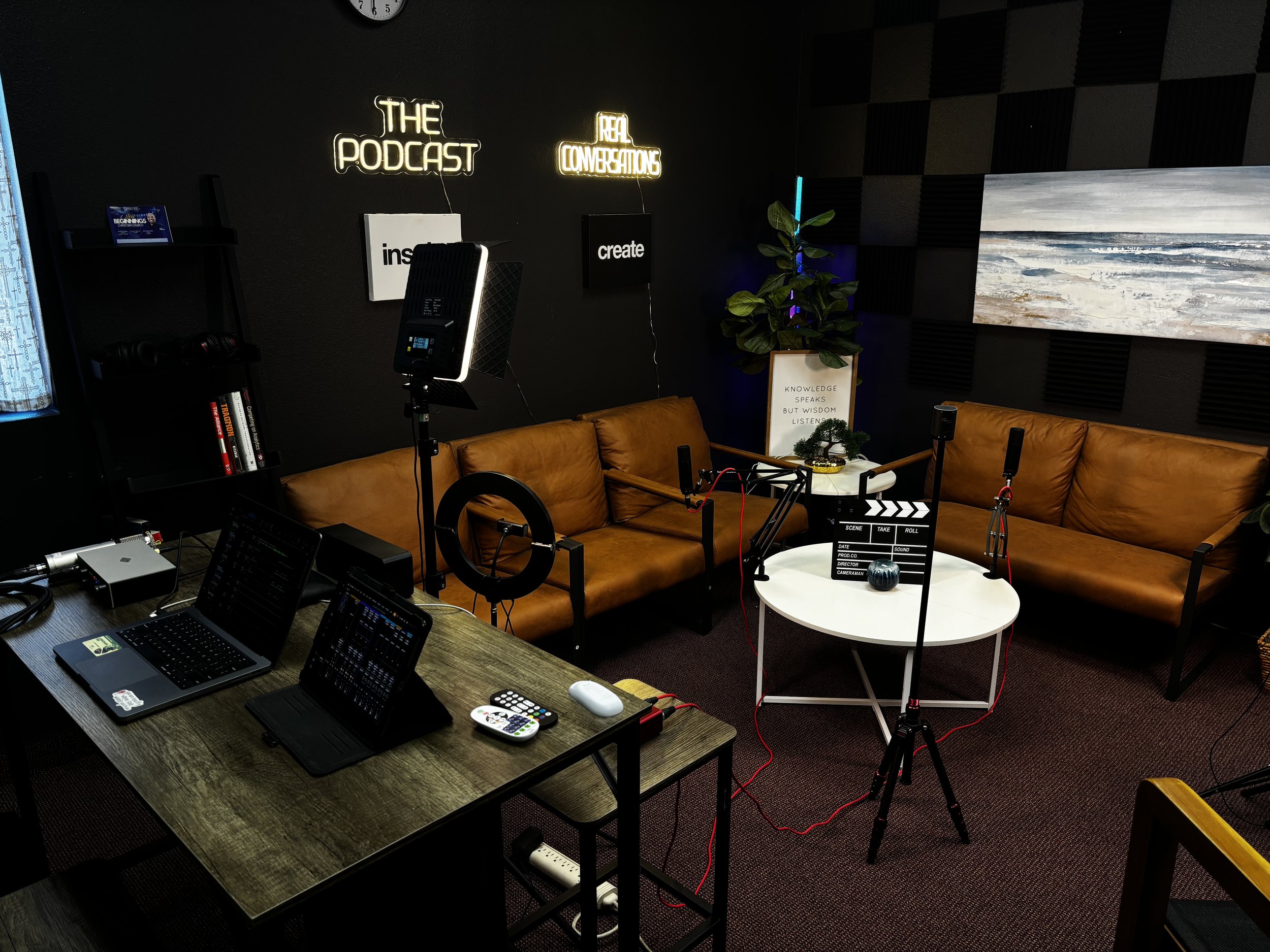 Podcast recording studio with a black wall, neon signs that say 'The Podcast' and 'Real Conversations,' a black bookshelf with books, a brown leather couch, a round white table with a decorative piece, cameras, microphones, ring light, laptop, and audio equipment.