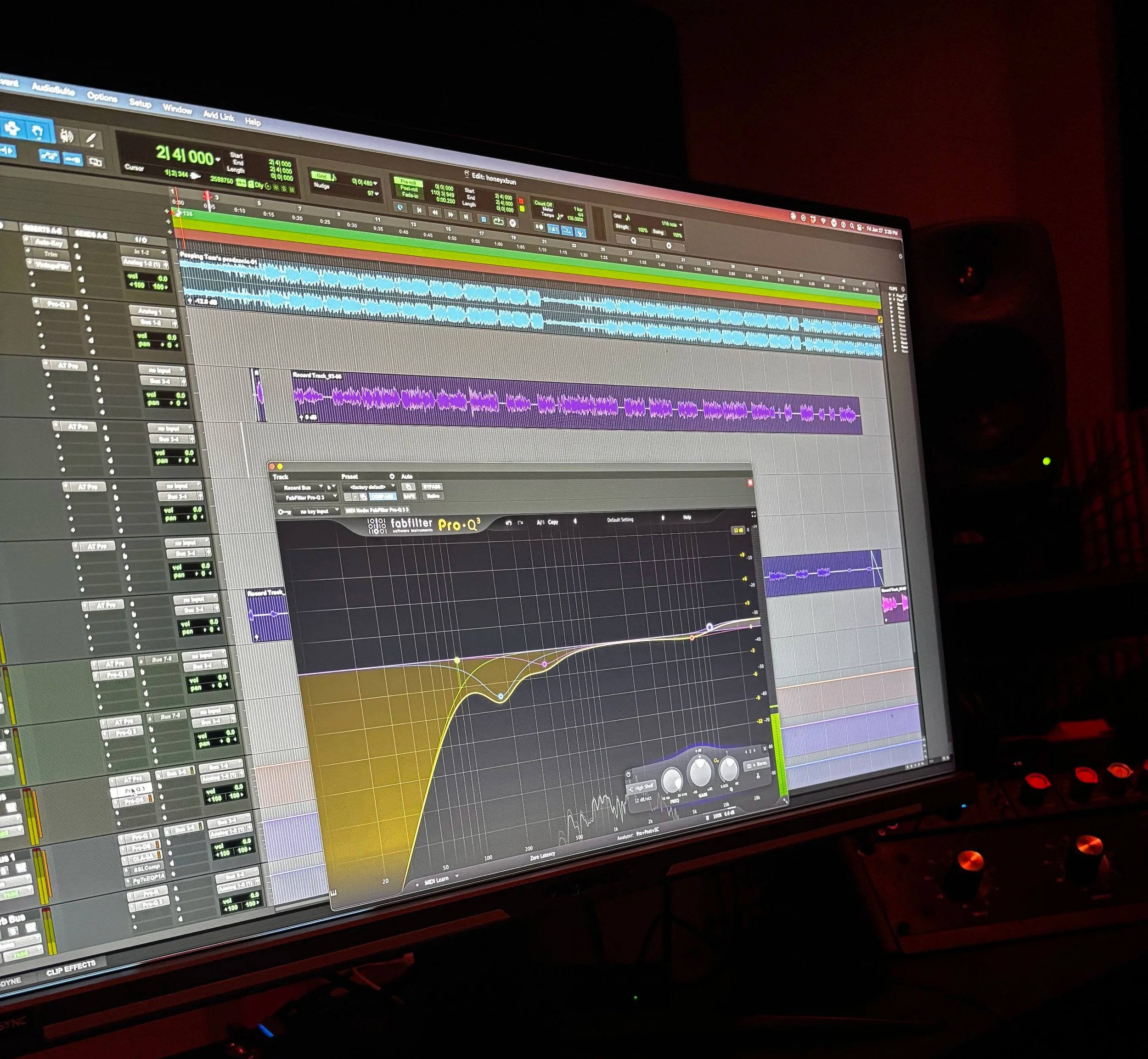 A computer monitor displaying digital audio workstation software with multiple tracks, waveforms, and an equalizer in a music studio environment.