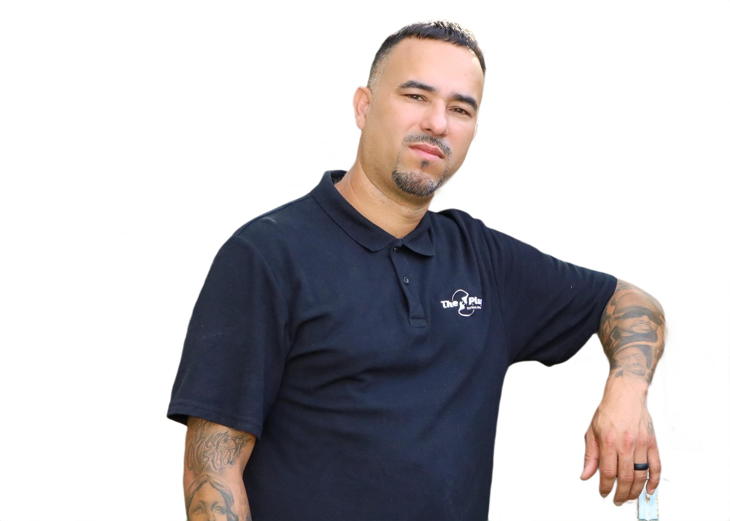 A man with tattoos on his arms wearing a black polo shirt, posing with his arm resting on a surface, looking at the camera with a neutral expression.