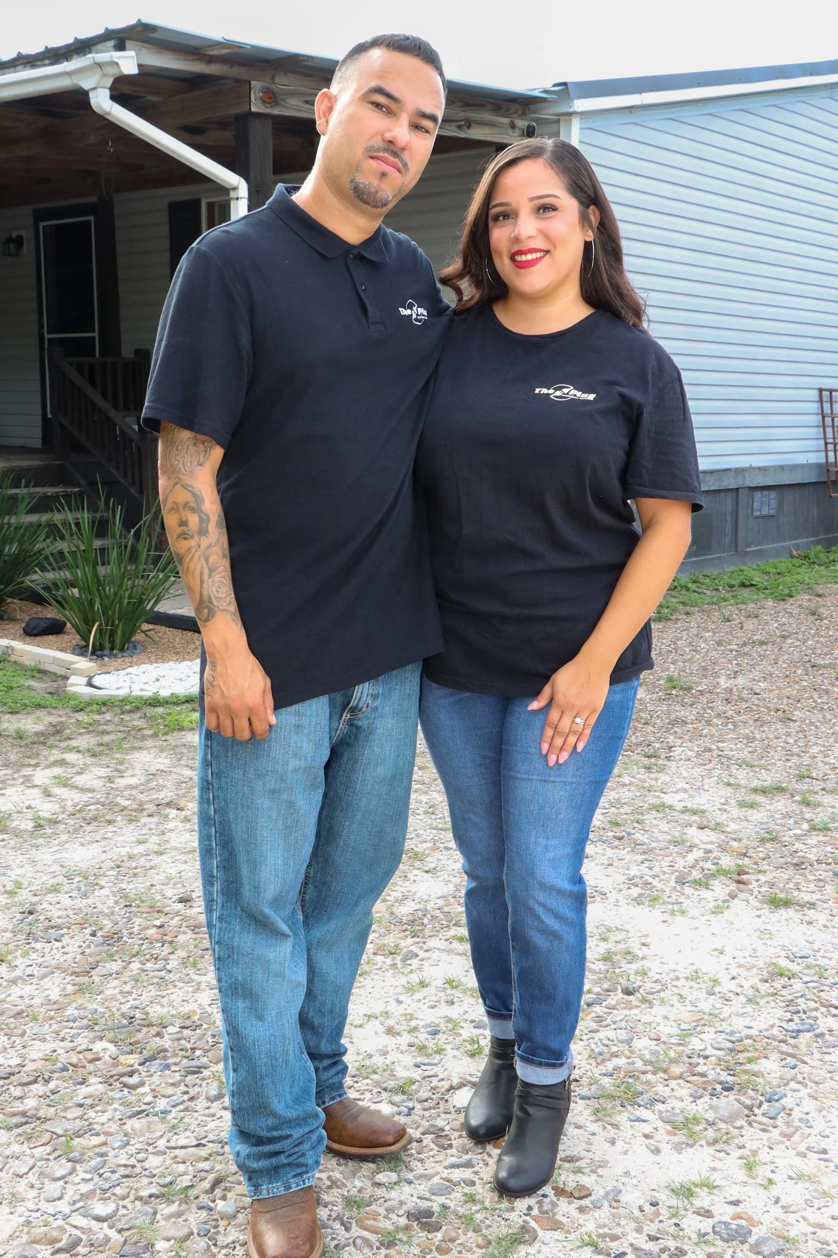 A man and woman standing outdoors in front of a house. The man has a goatee and tattoos on his arm, wearing a black polo shirt and jeans. The woman has shoulder-length dark hair, red lipstick, and hoop earrings, wearing a black T-shirt and jeans. They are smiling and posing closely.