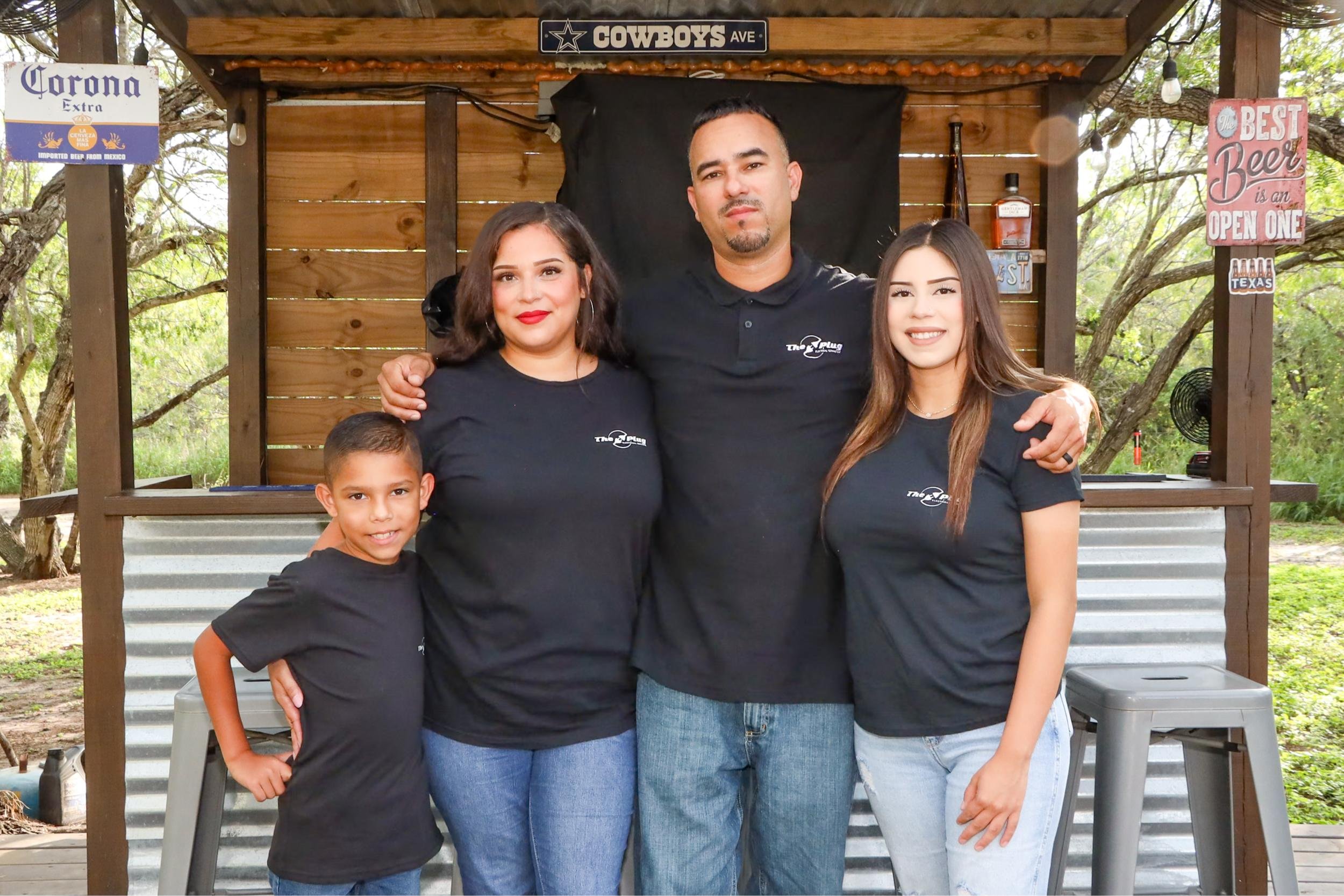 A family of four standing together outdoors in front of a wooden bar with signs, wearing matching black shirts. The woman has shoulder-length dark hair and red lipstick, the man has short dark hair and a goatee, and the two children, a boy and a girl, are smiling. The background shows trees and greenery.