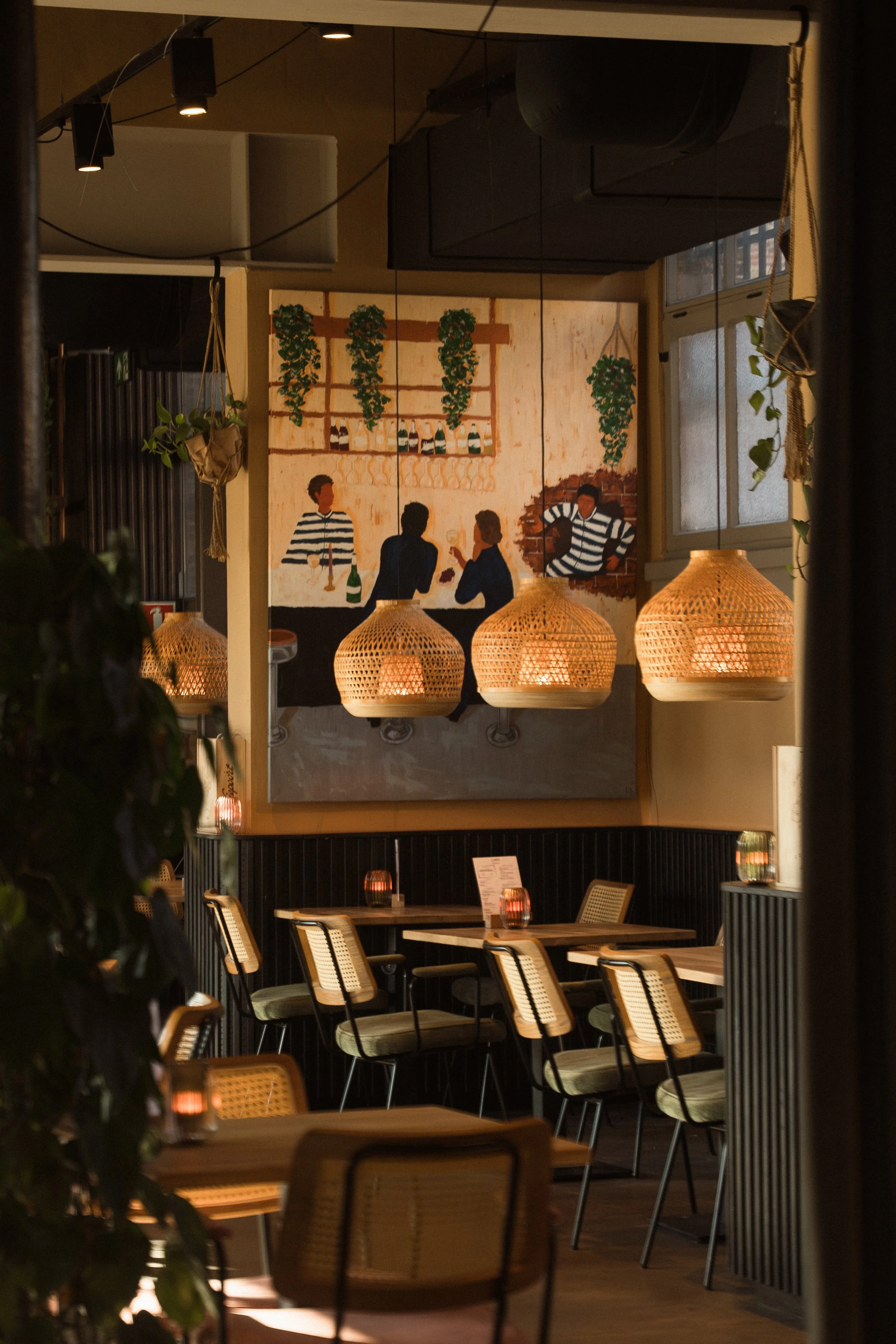 Interieur van een restaurant met geblazen glazen hanglampen, een grote muurkunstwerk met mensen die aan de bar zitten, en gezellige tafels versierd met kaarsen.