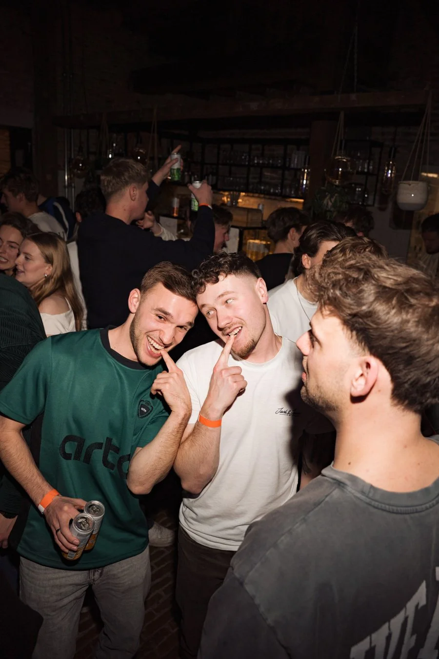 Drie jonge mannen op een feest of bar, glimlachend en vinger tegen hun neus, omgeven door andere feestvierders.
