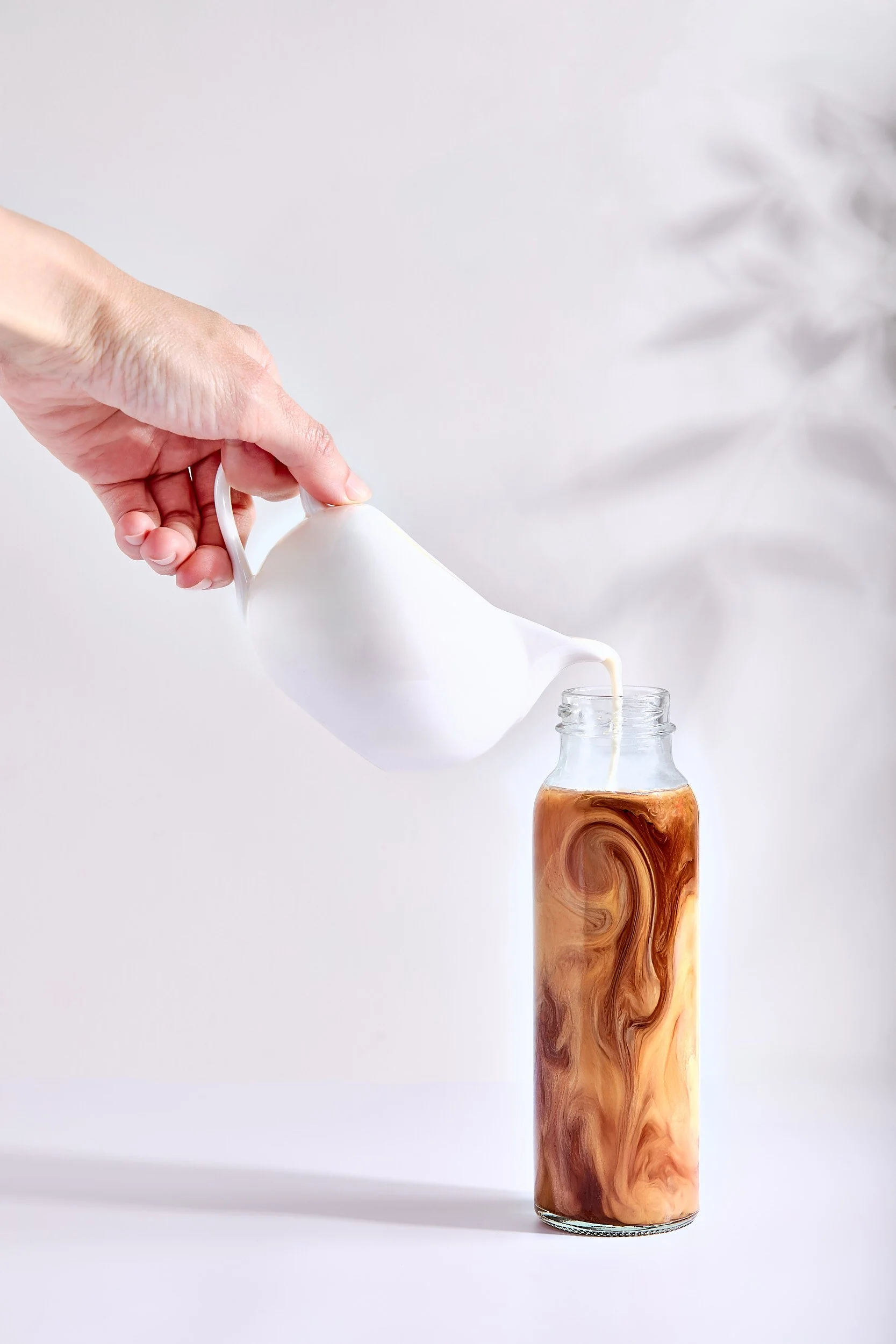 Hand pouring creamer into a jar of iced coffee with swirled milk design. Light and bright drink photography, beverage photo.