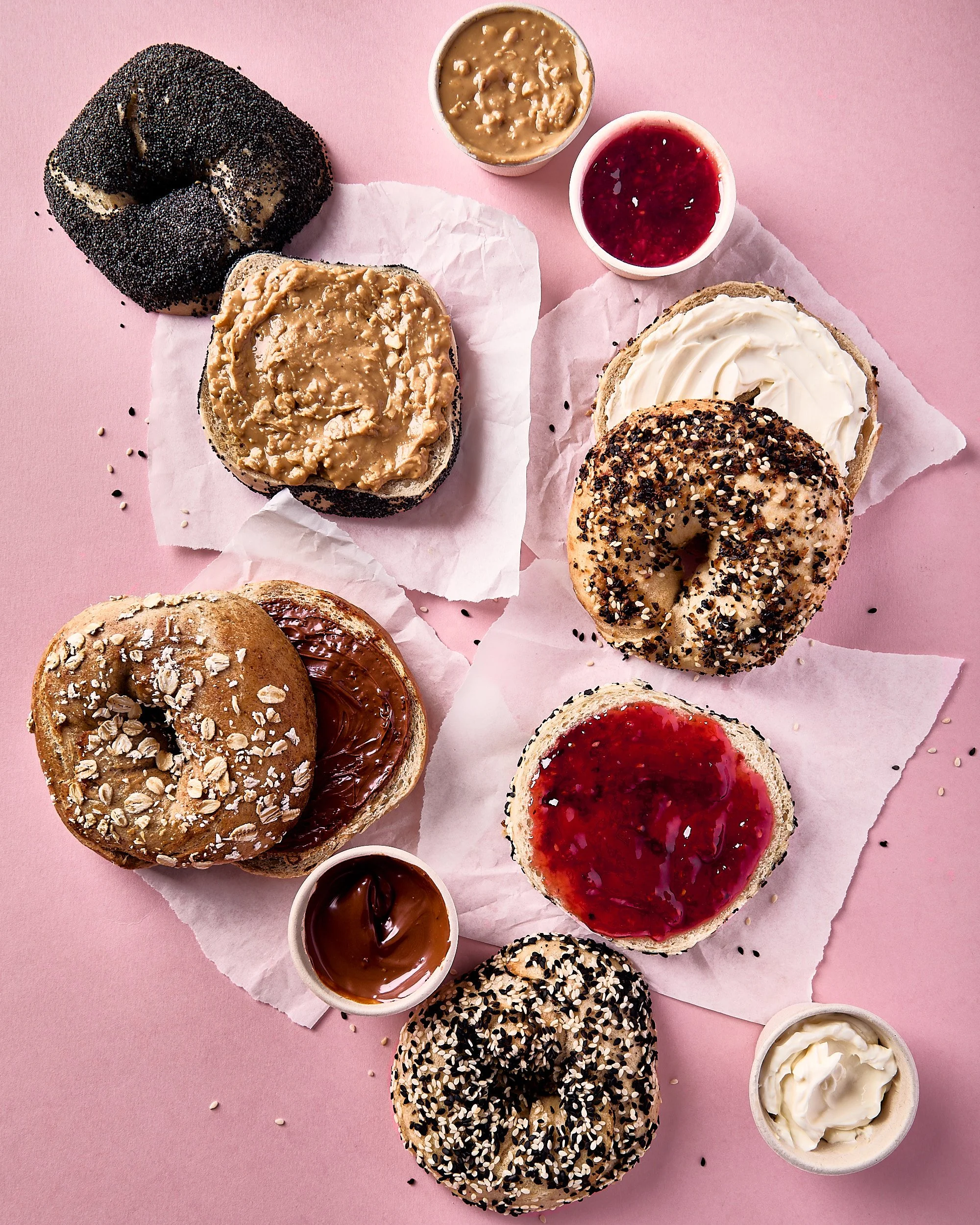 Assorted donuts topped with various glazes and sprinkles, along with small cups of jam, chocolate spread, and cream, arranged on pink parchment paper and a pink background. Cafe photography, brunch food photography.