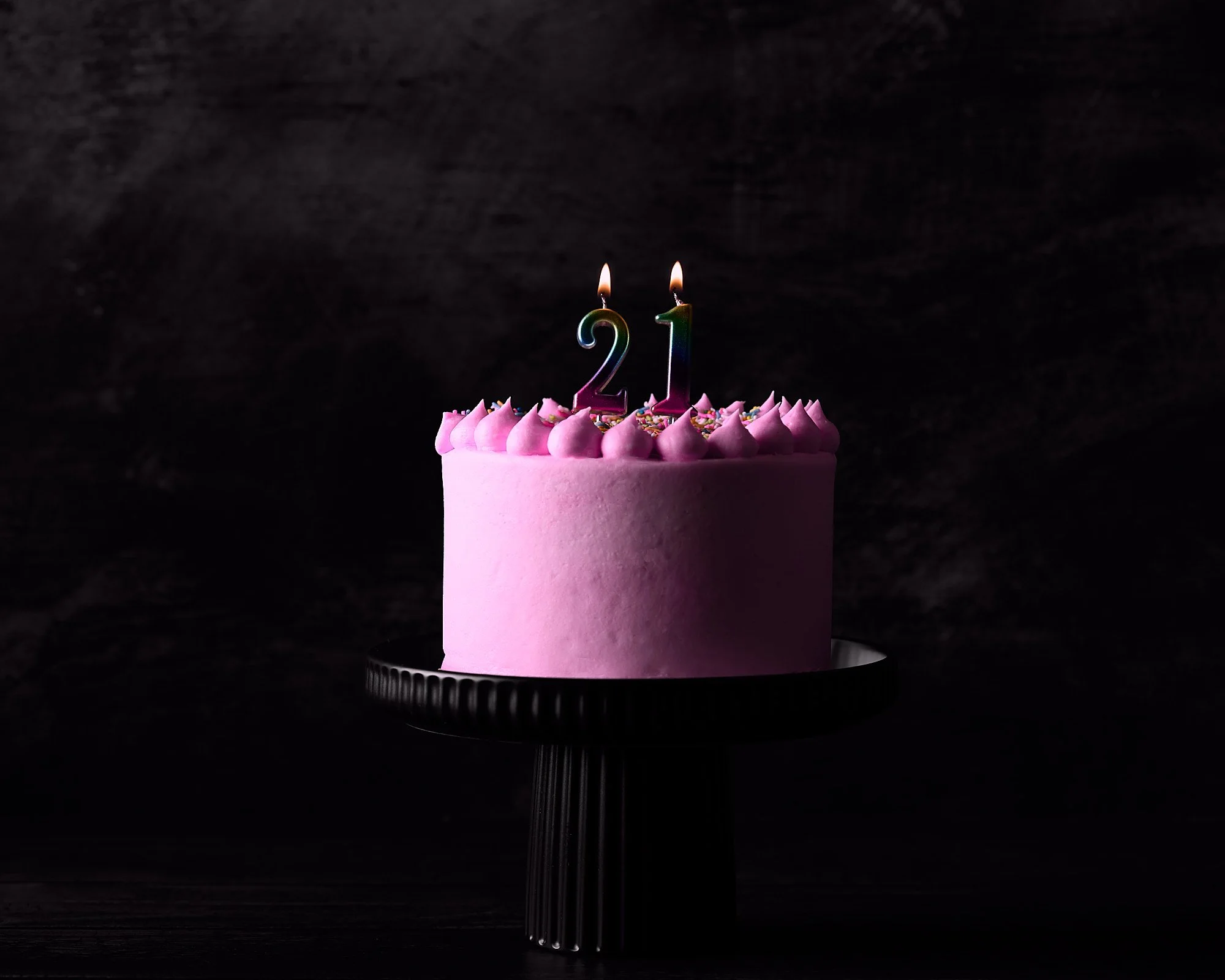 Pink birthday cake with two lit candles shaped as the numbers 2 and 1, decorated with pink frosting swirls and colorful sprinkles, on a black pedestal against a dark background. Dessert photo, dessert photography in Sydney