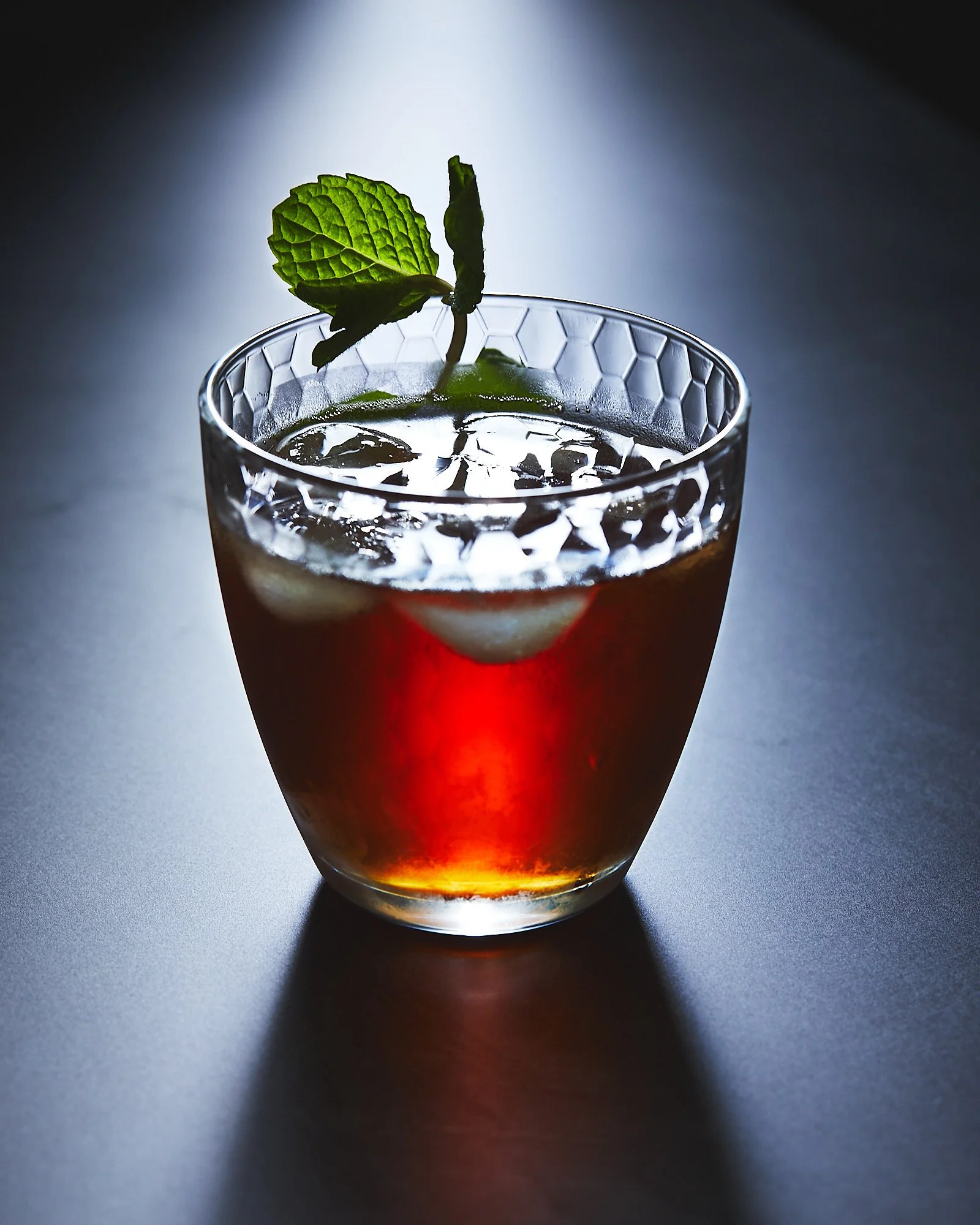 A glass of dark-colored cocktail with ice cubes and a mint leaf garnish on top. Drink photography, beverage photographer 