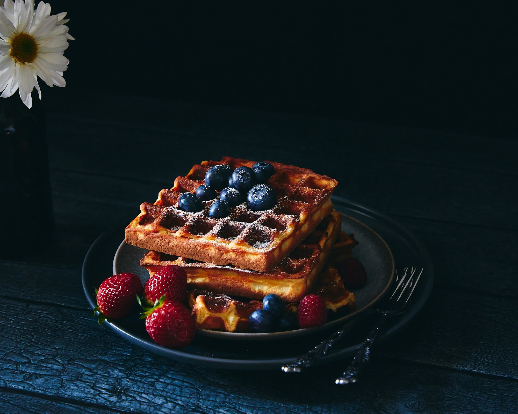 Stacked waffles topped with blueberries and dusted with powdered sugar, served with strawberries on a black plate, on a dark wooden table. Dark and moody food photography, dessert photography, brunch photographer. 