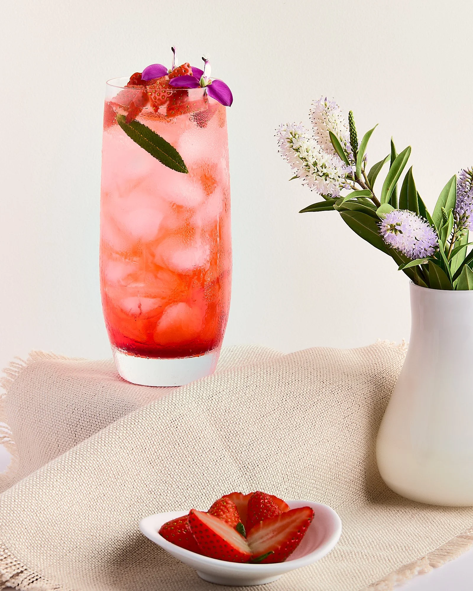 A tall glass of pink strawberry lemonade with ice, garnished with strawberry slices and purple flowers, placed on a beige cloth next to a white vase with purple and green flowers, and a small dish of halved strawberries. Sydney drink photographer. 