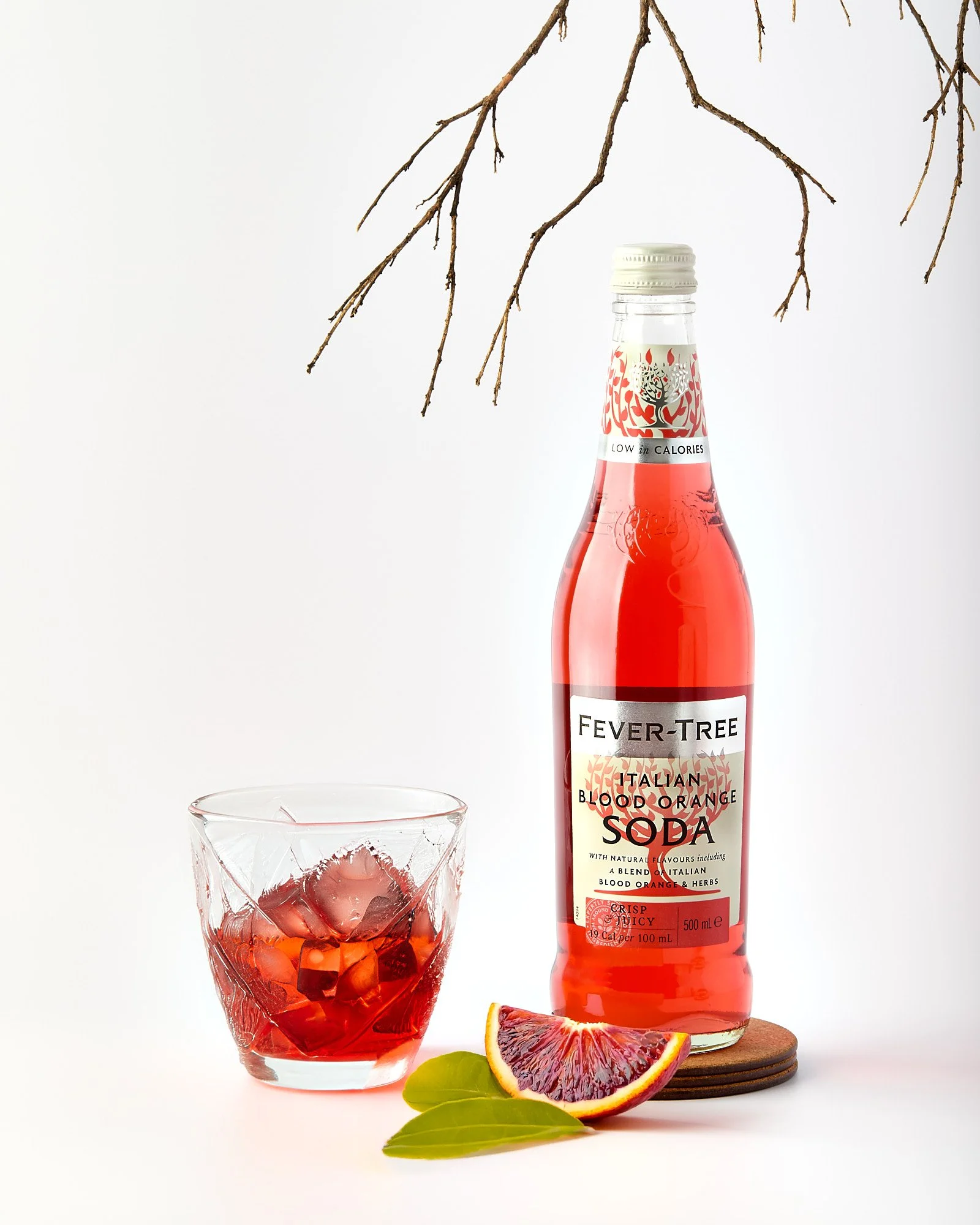 Bottle of Fever-Tree Italian Blood Orange Soda with a glass filled with ice and soda, a wedge of blood orange, and some green leaves on a white surface with a branch hanging above. Commercial, brand photography