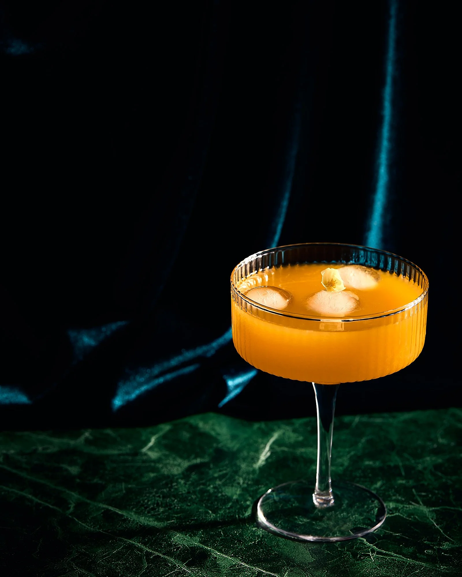 A yellow cocktail with ice cubes and a small garnish in a clear, textured cocktail glass on a green leaf-patterned surface against a dark background. Beverage photographer, drink photography, photo taken by Mah Vafaei in Sydney Australia.